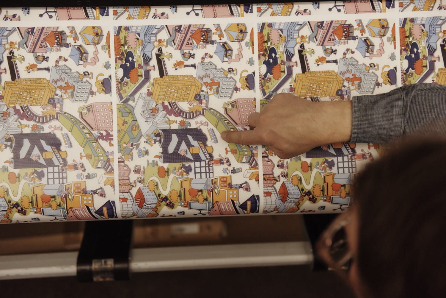 A person points to a colorful, illustrated map being printed, featuring various buildings, people, and city scenes in a cartoon style—perfect for learning how to transfer vibrant designs onto posters. Another person is partially visible in the foreground.