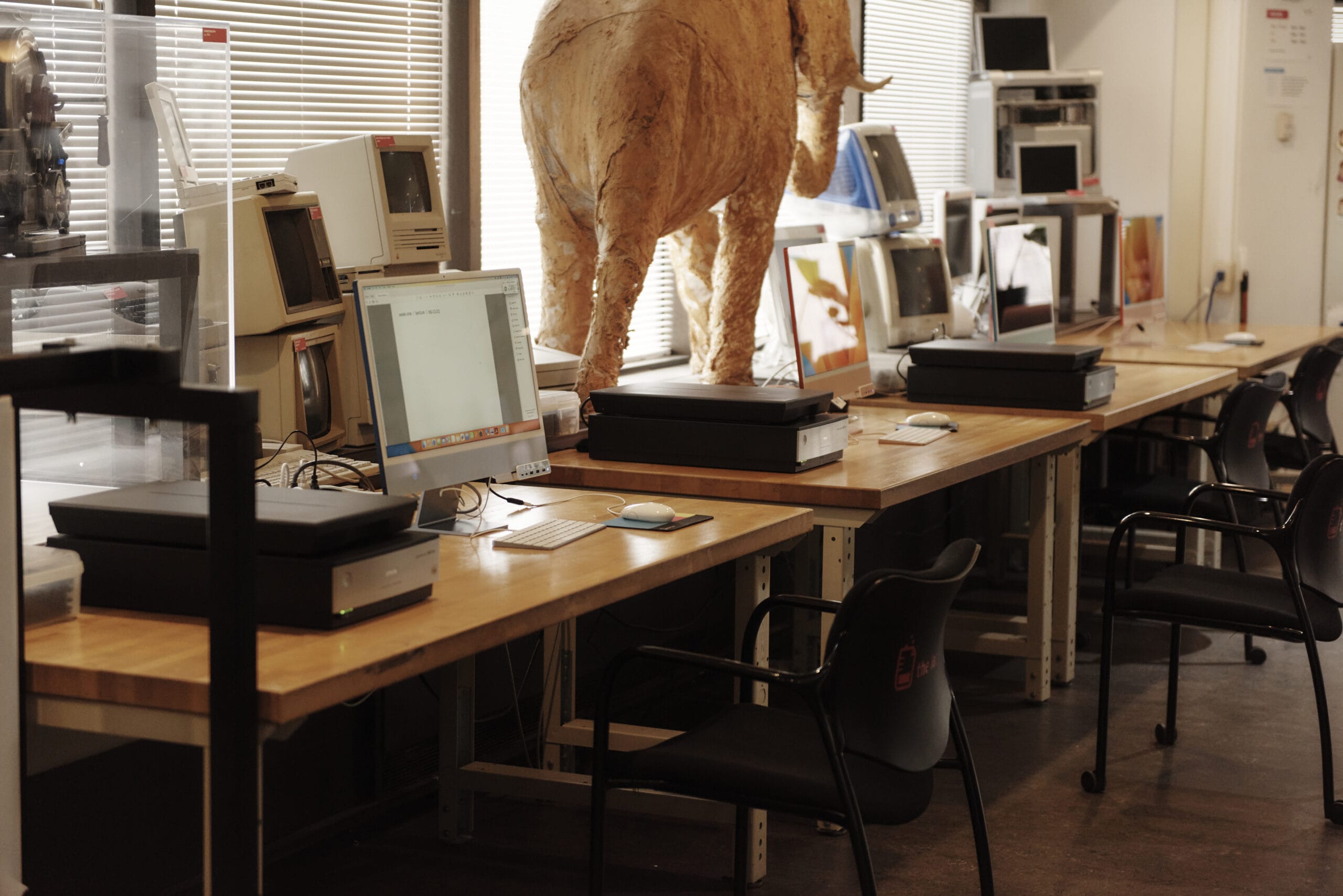 A workspace with wooden tables featuring computers, scanners, and various electronics. An elephant sculpture made of light-colored material stands on one table. Chairs are arranged in front of the workstations.