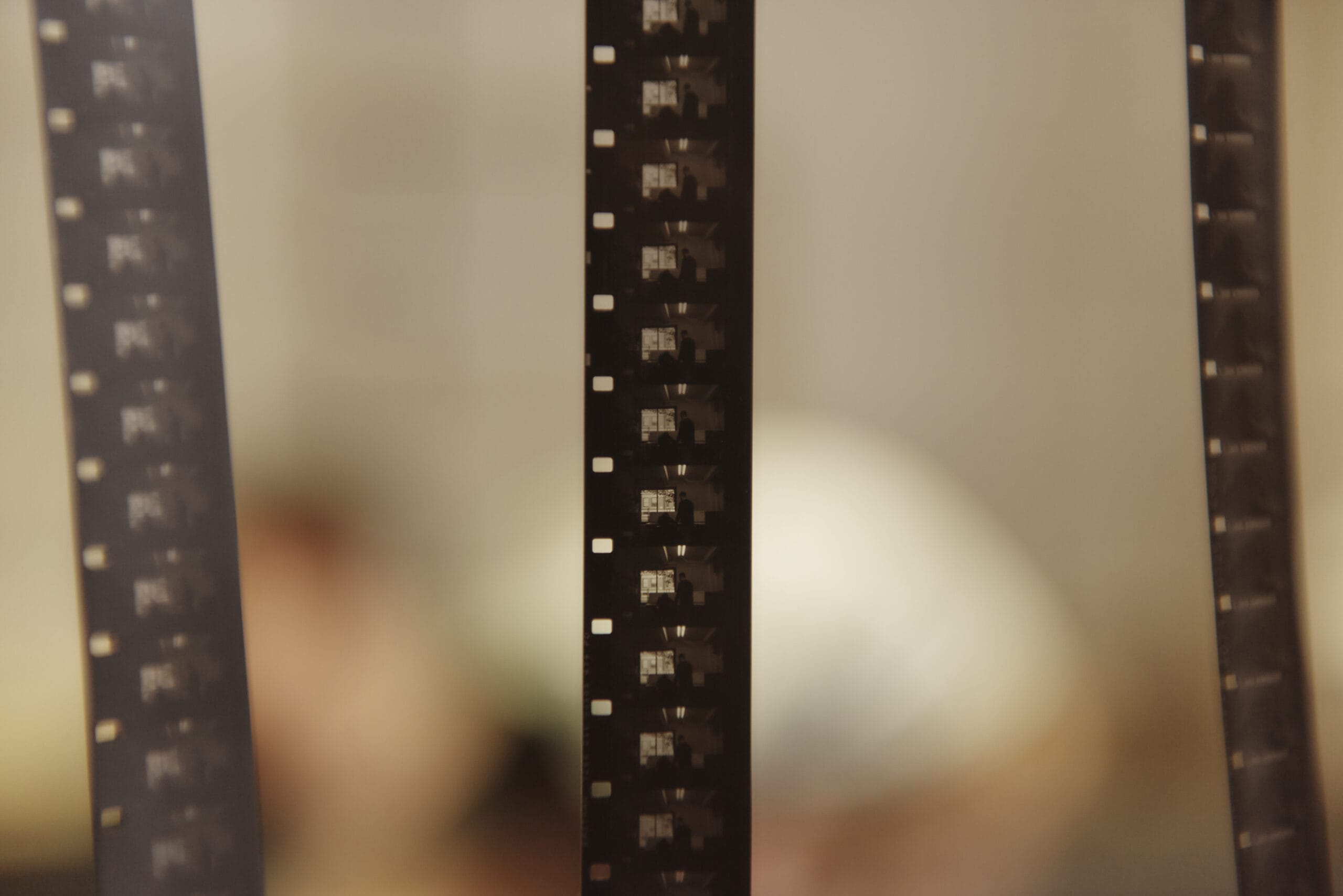 Close-up of several strips of photographic film hanging vertically. The background is blurred, showing indistinct shapes and colors, creating a soft and artistic feel.
