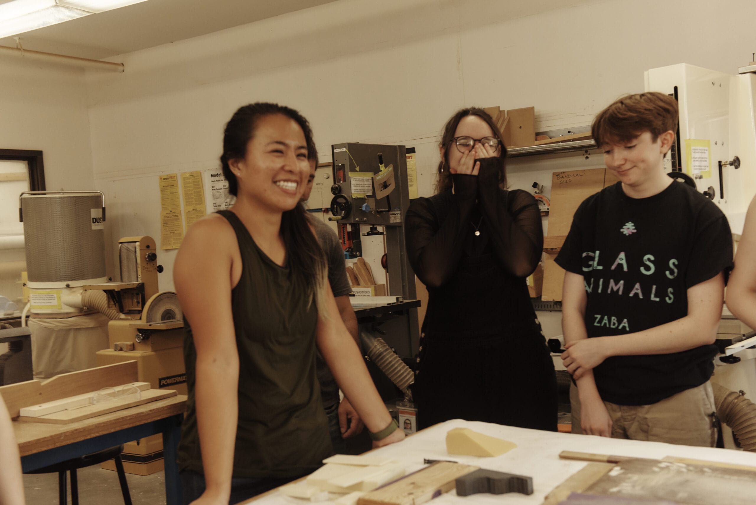 Three people stand around a worktable in a workshop, smiling and laughing. Surrounded by tools and wood pieces, they collaborate to aid each other with their projects, making creative work enjoyable while discussing ways to manage costs together.