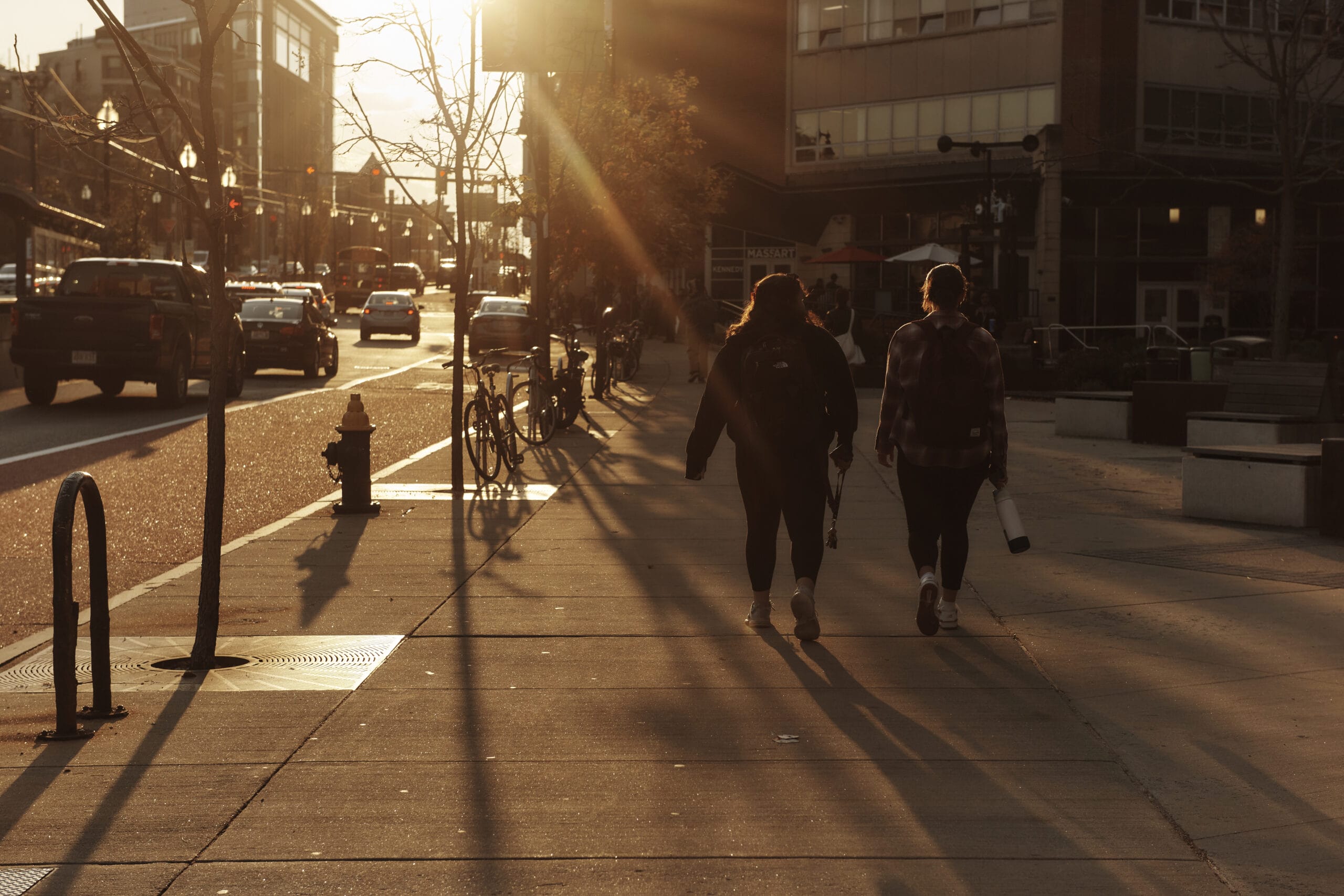 Two people walk on a sunlit city sidewalk, casting long shadows. Bicycles are parked nearby, and cars line the street. As they discuss the transfer process, the warm glow of the setting sun adds a serene atmosphere to the urban scene.
