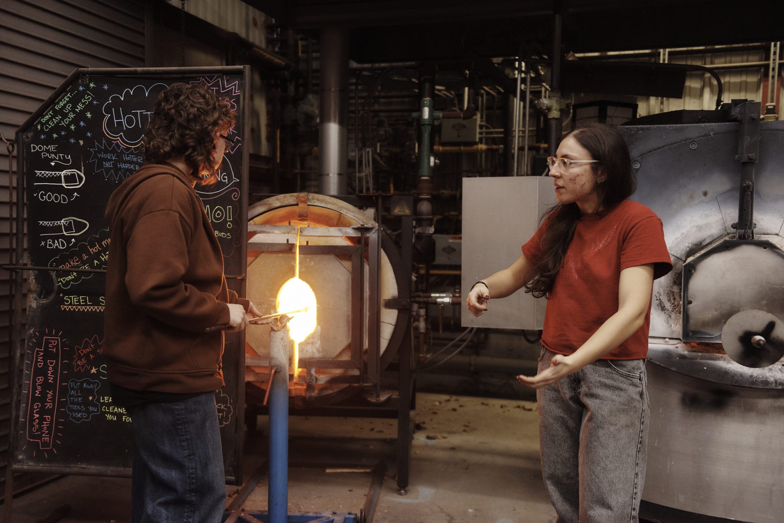 Two people engaging in glassblowing in an MFA studio. One skillfully shapes molten glass with precision tools while the other gestures meaningfully, possibly discussing 3D Fine Arts techniques. A blackboard with colorful notes and industrial equipment adds depth to their creative space.
