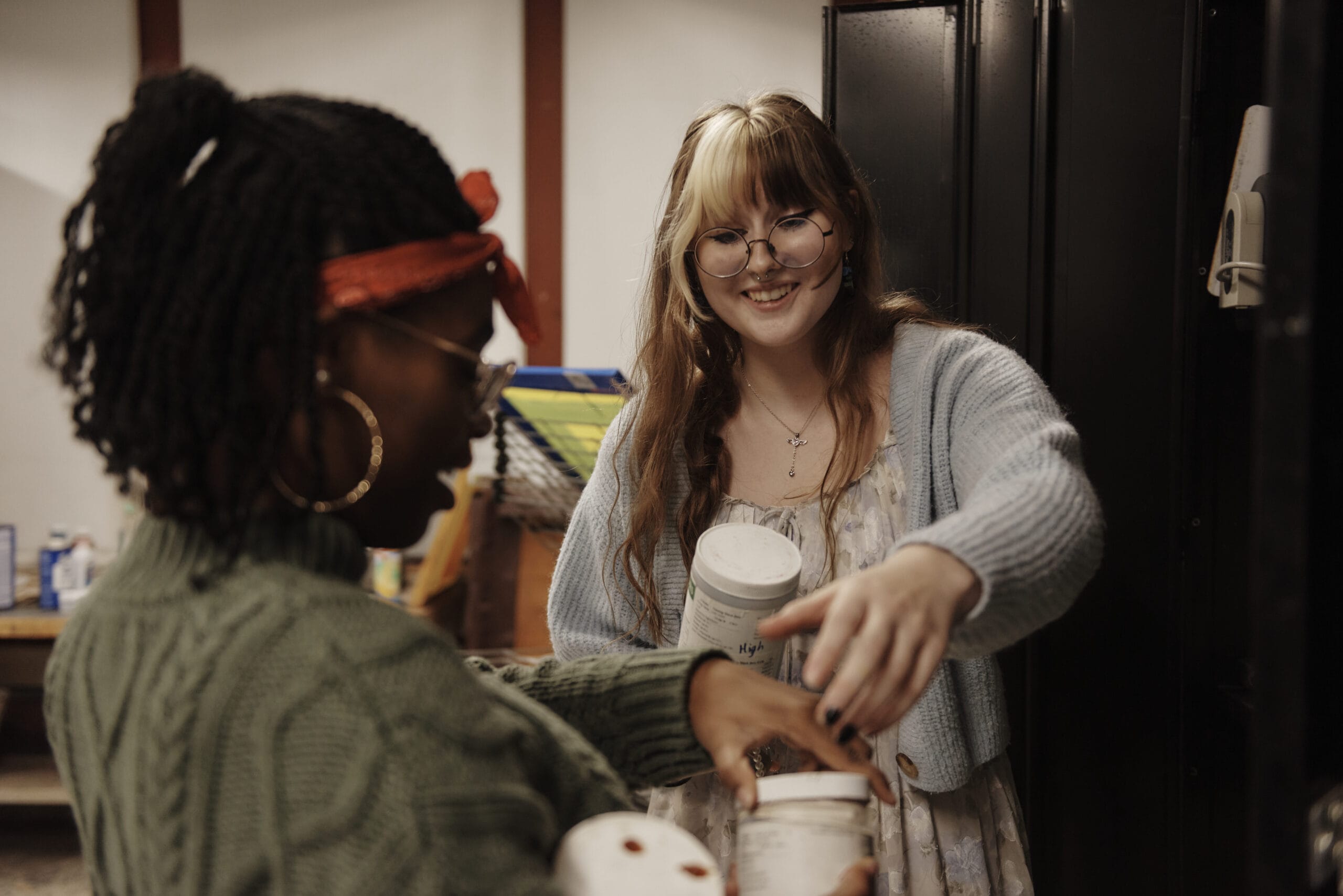 Two people smile and hold jars in a room. One, wearing glasses and a cardigan, gestures while the other sports a green sweater, red headband, and hoop earrings. It’s an atmosphere of student accessibility as they engage in their shared project with enthusiasm. A cabinet is visible in the background.