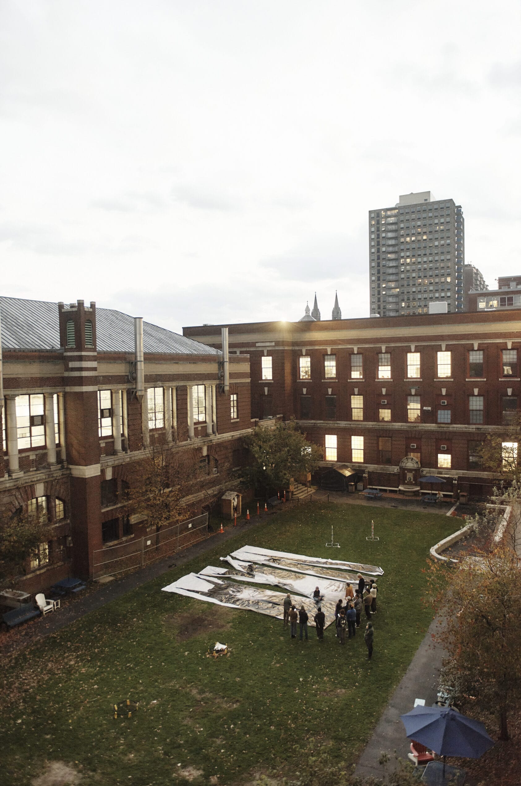 An aerial view shows students gathered around large, unrolled canvases on a grassy courtyard surrounded by brick buildings. The scene captures the essence of education under overcast skies, with a tall building standing prominently in the background.