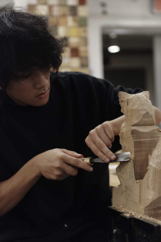 A person with dark hair is intently carving a wooden sculpture using a chisel in a workshop, meeting their artistic admissions application deadlines. The background features a wall with a multicolored tile pattern, echoing the vibrancy and dedication of the craftsman.