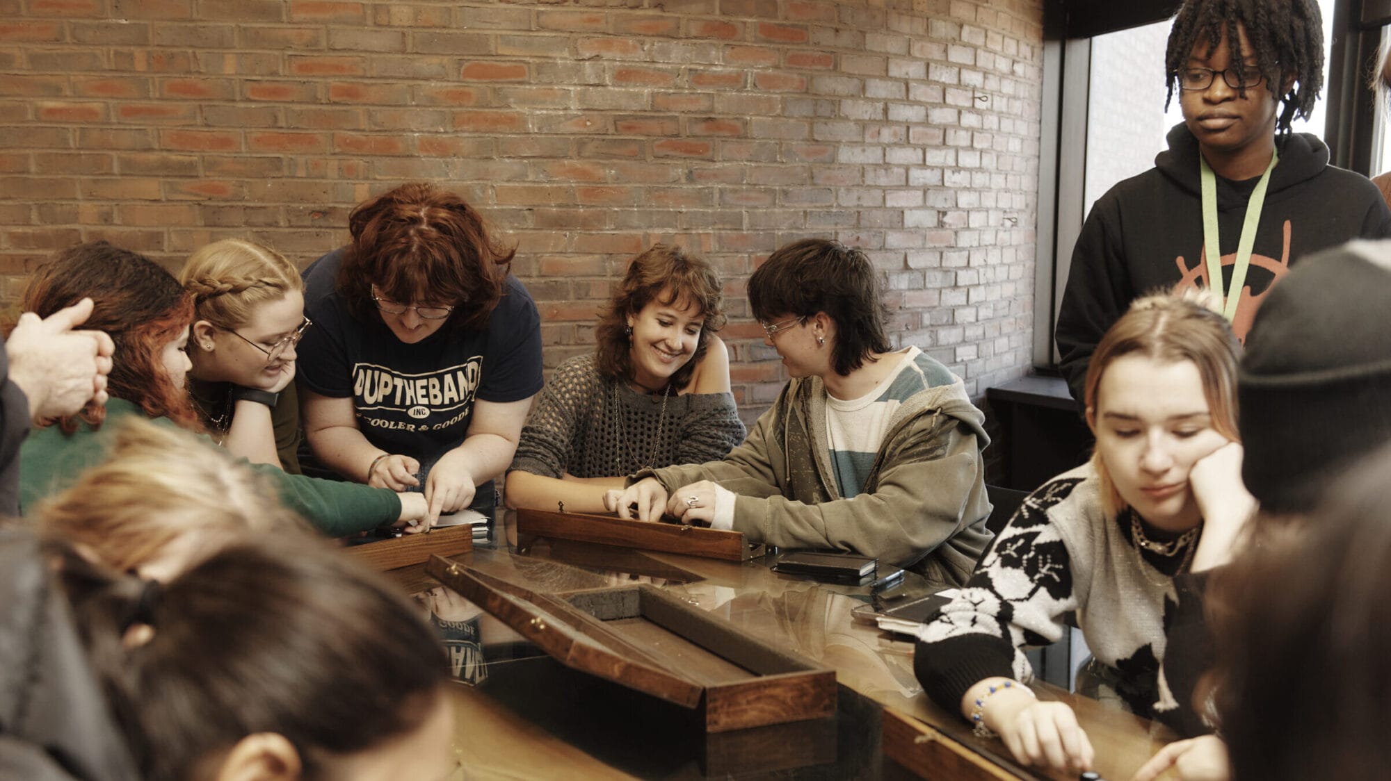 A group of people sitting around a table playing a board game. The room has brick walls and large windows. Some individuals are smiling and engaged in the game, while others are observing or contemplating their moves.