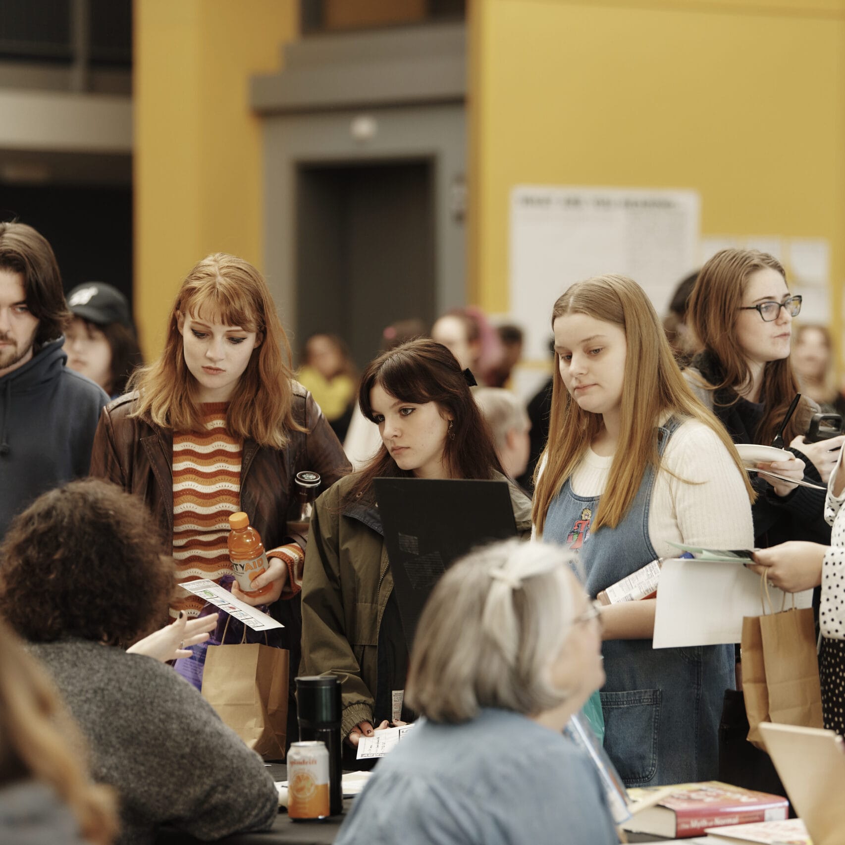 Students stand around a table listening to a staff member speak, at a crowded student fair.