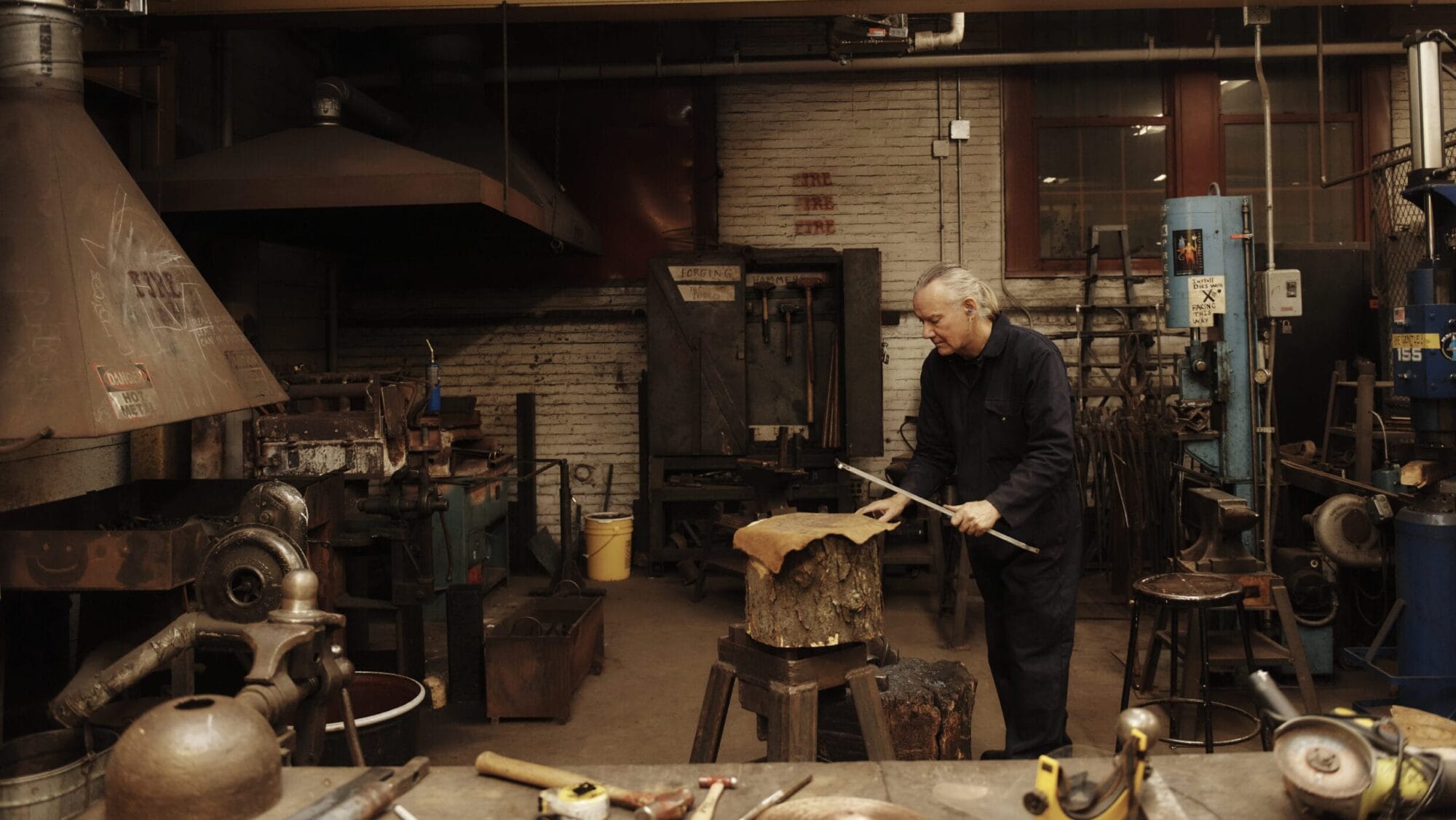A person wearing a dark jumpsuit works with metal tools in a cluttered workshop. The space resembles a 3D Fine Arts studio, filled with equipment like anvils, grinders, and hammers. The person is an MFA student focused on perfecting a metal piece resting on a wooden stump.
