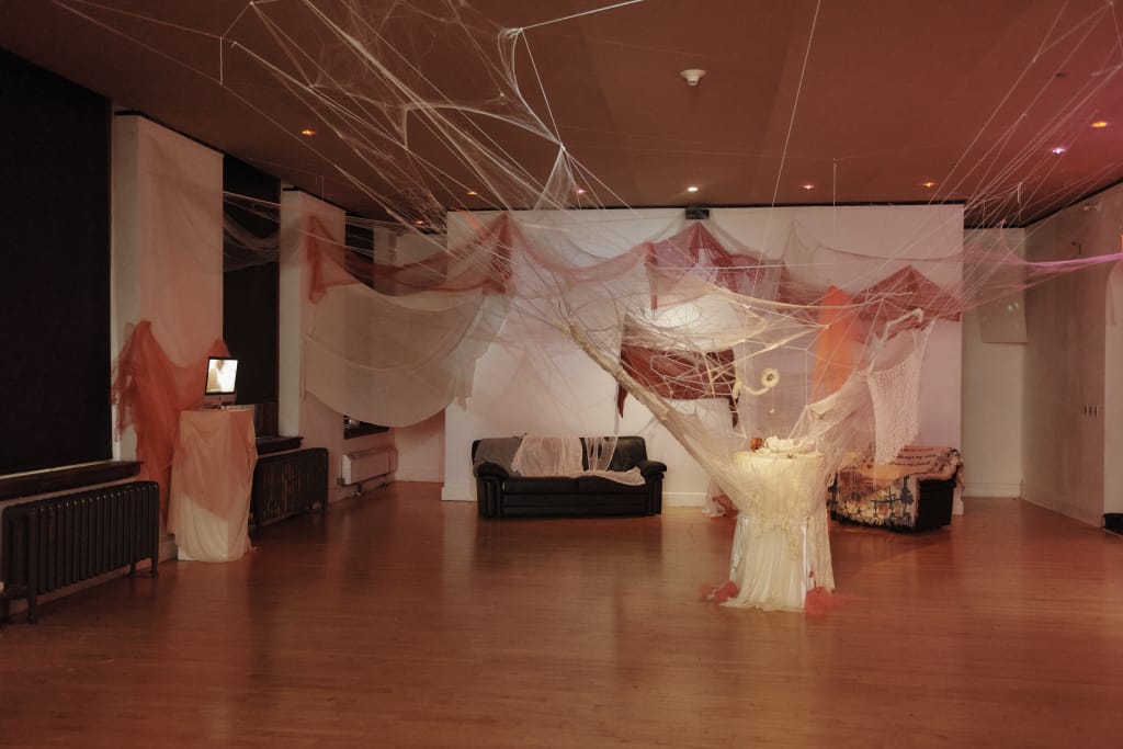 The room, reminiscent of avant-garde art galleries, features extensive spider web-like decorations on the ceiling and walls. A couch and table draped with fabric occupy the center. Soft lighting enhances the spaces mysterious, atmospheric feel, akin to an intriguing exhibition at MassArt.