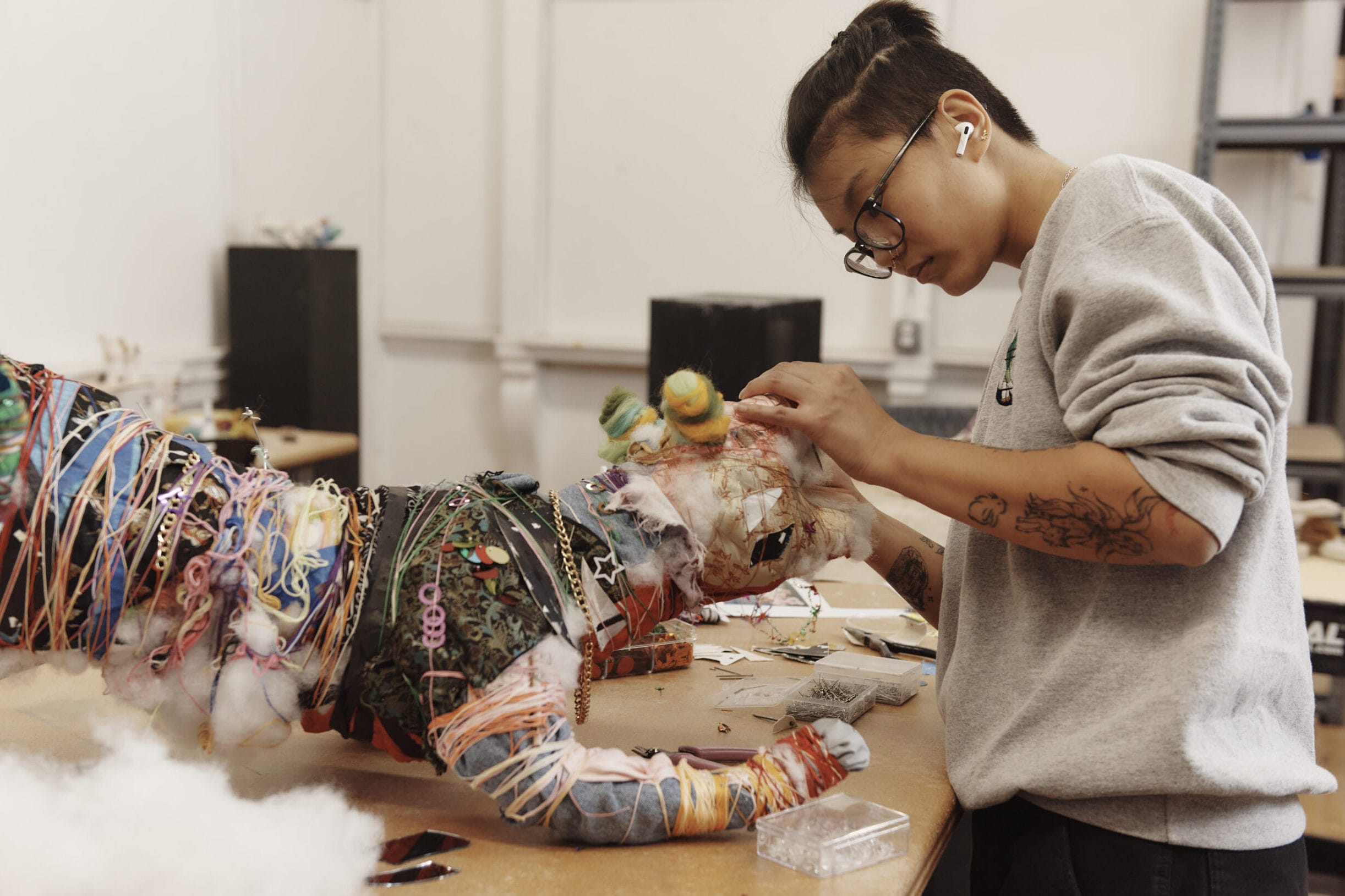 A person wearing glasses and a sweater is intricately working on a colorful, textured sculpture of a creature on a desk in a workshop. The sculpture is made from various materials like yarn and fabric. Shelves and tools are visible in the background.