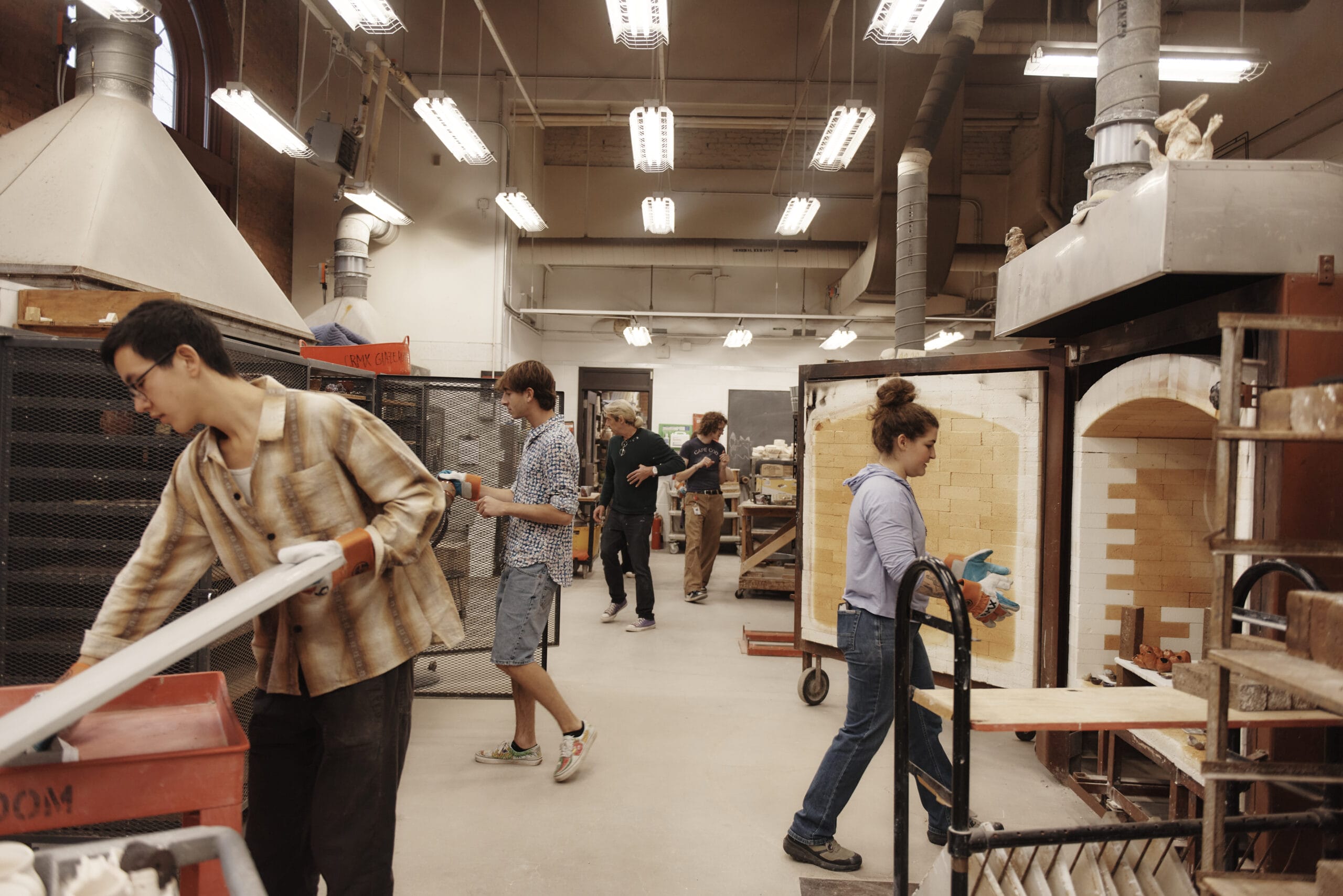 A group of people are working in a pottery studio filled with kilns, pottery wheels, and tools. One person is carrying a ceramic piece, while others are busy with various tasks. The room is well-lit with overhead lights.