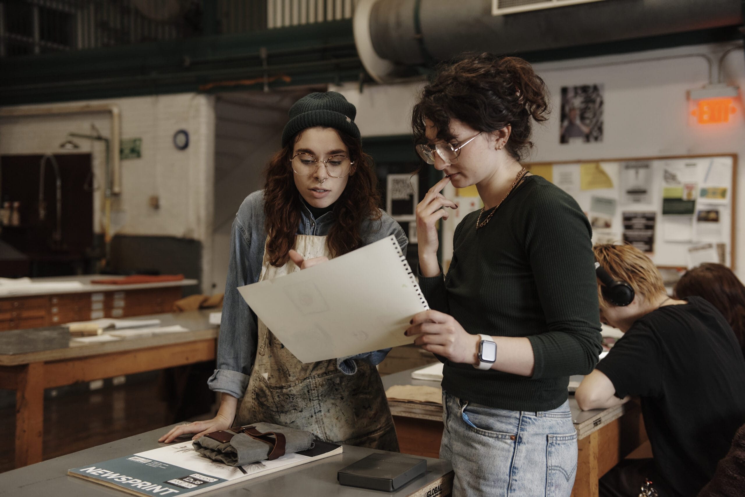 Two people in a workshop discuss a paper design. One wears a beanie and apron, the other glasses and a smartwatch. The background includes tables, art materials, and a bulletin board. Others are working at tables.