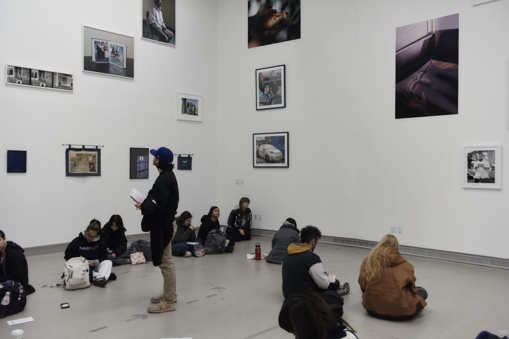 A group of people sit on the floor or stand inside a gallery room with white walls. Various framed photographs are displayed on the walls. One person stands, appearing to take notes.