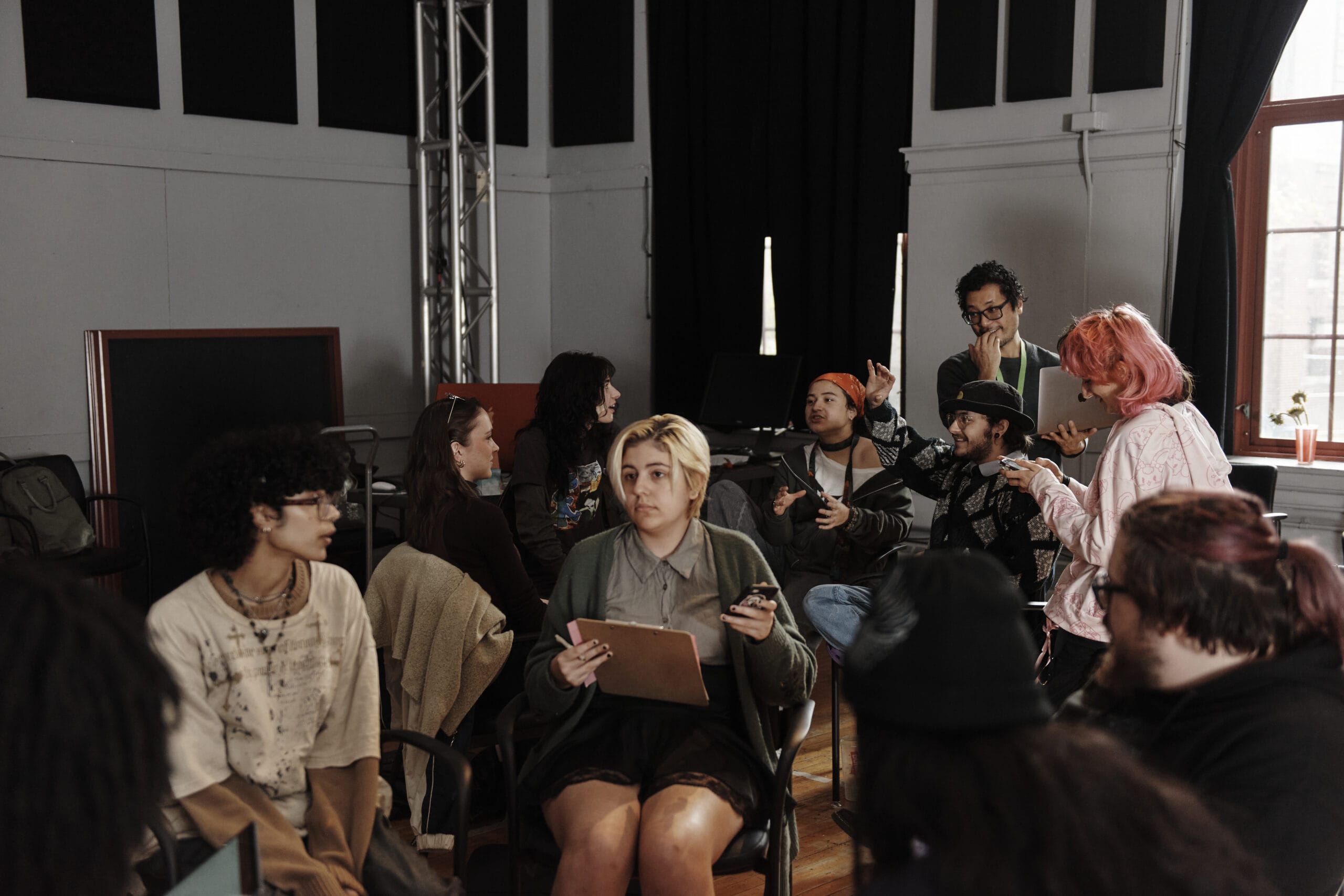 A group of people, gathered in a circle inside the room with dark curtains and large windows, appear deeply engaged in discussing admissions application deadlines. Some hold notepads and phones, while they converse over the guiding dates on the wooden floor.