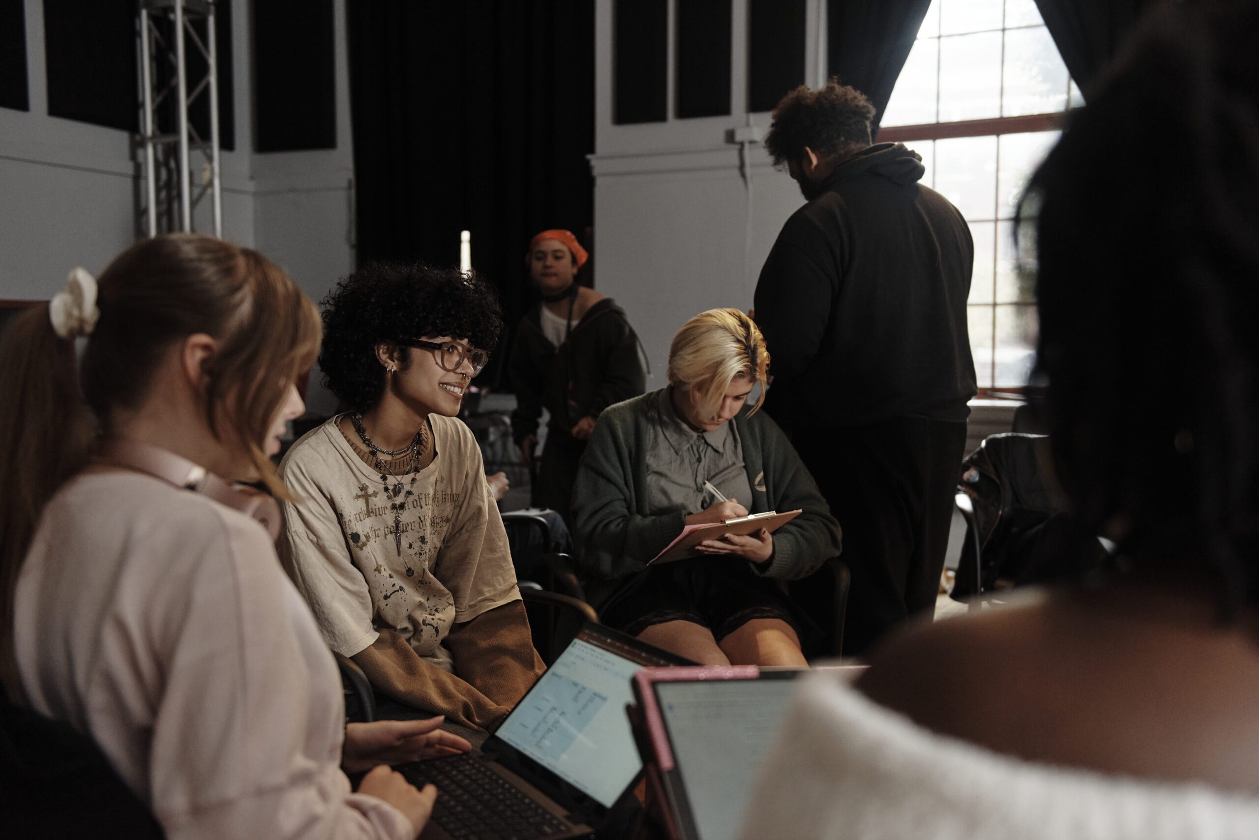A group of people are sitting and standing in a room, some with laptops and tablets. They appear to be working or studying together. The environment looks casual and collaborative, with a focused atmosphere.