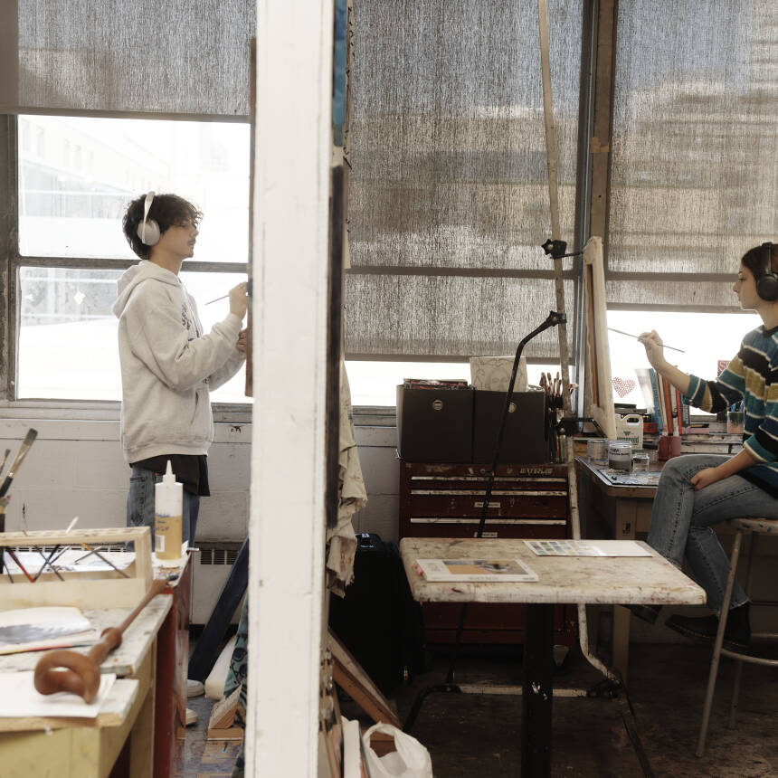 The side view of a wall divides a photo of two students painting in their respective studio spaces.