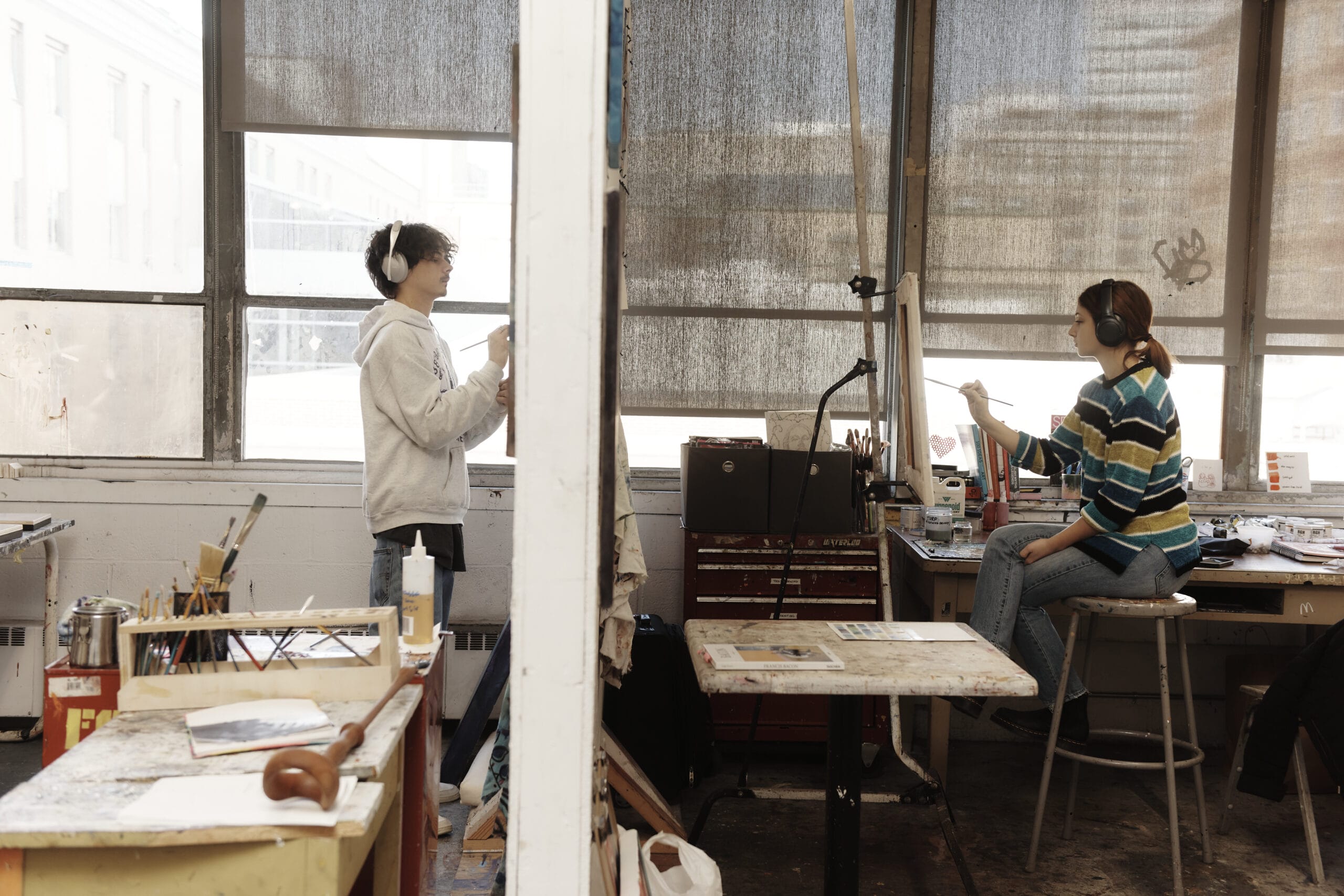 The side view of a wall divides a photo of two students painting in their respective studio spaces.