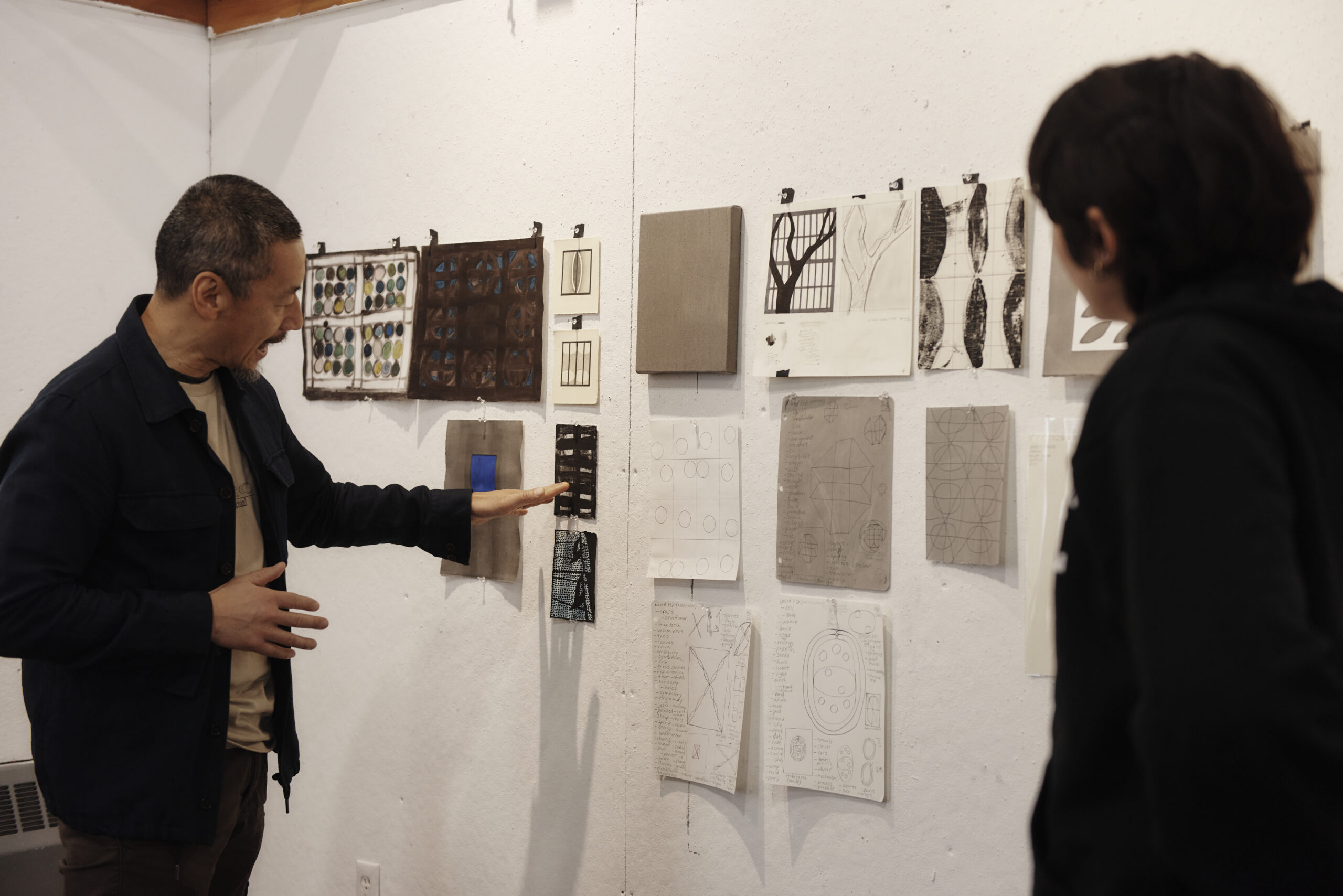 A professor points and talks about a student work hanging up on a while while a student observes and listens.