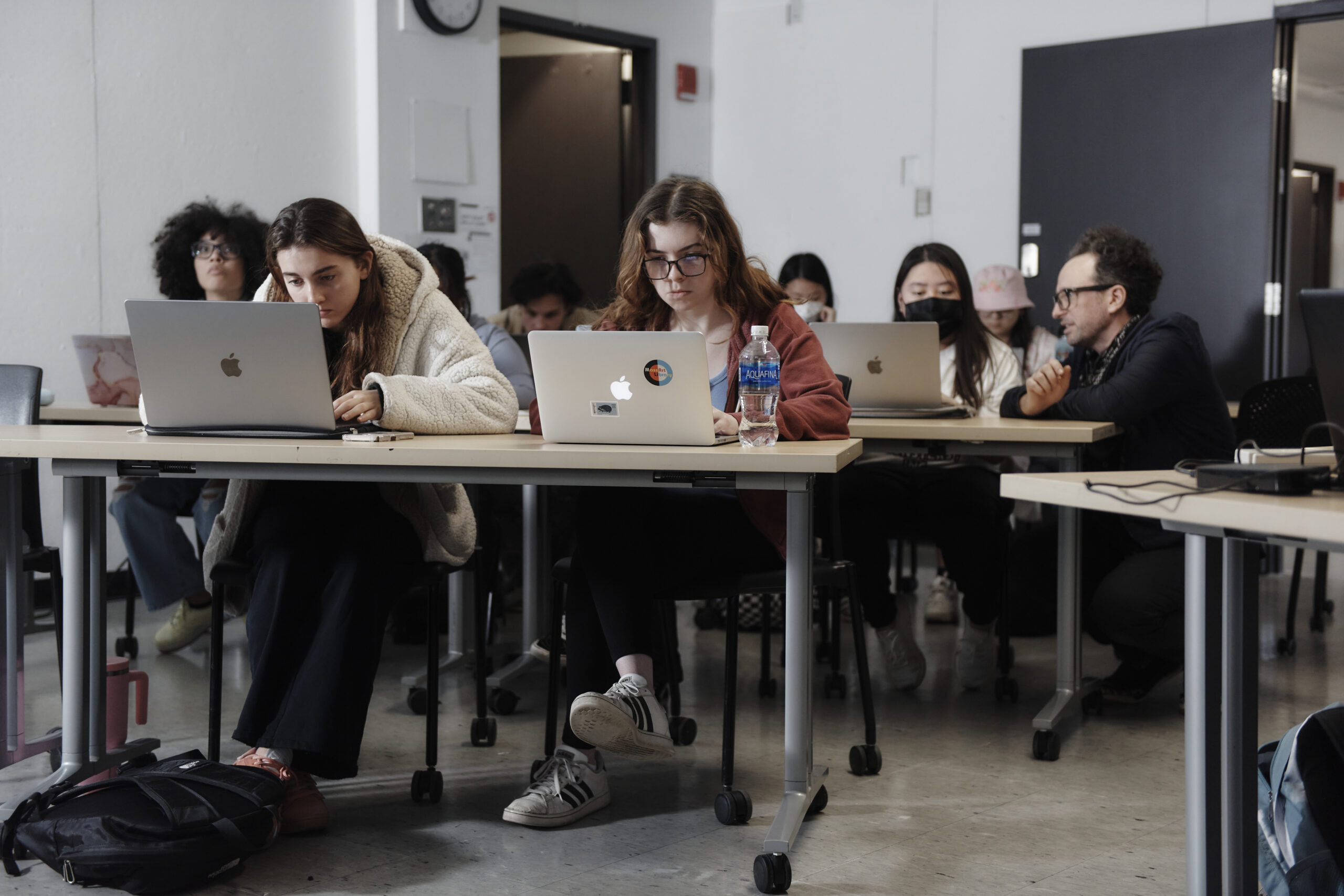 Students sit staring at their laptops at rows of tables in a classroom.