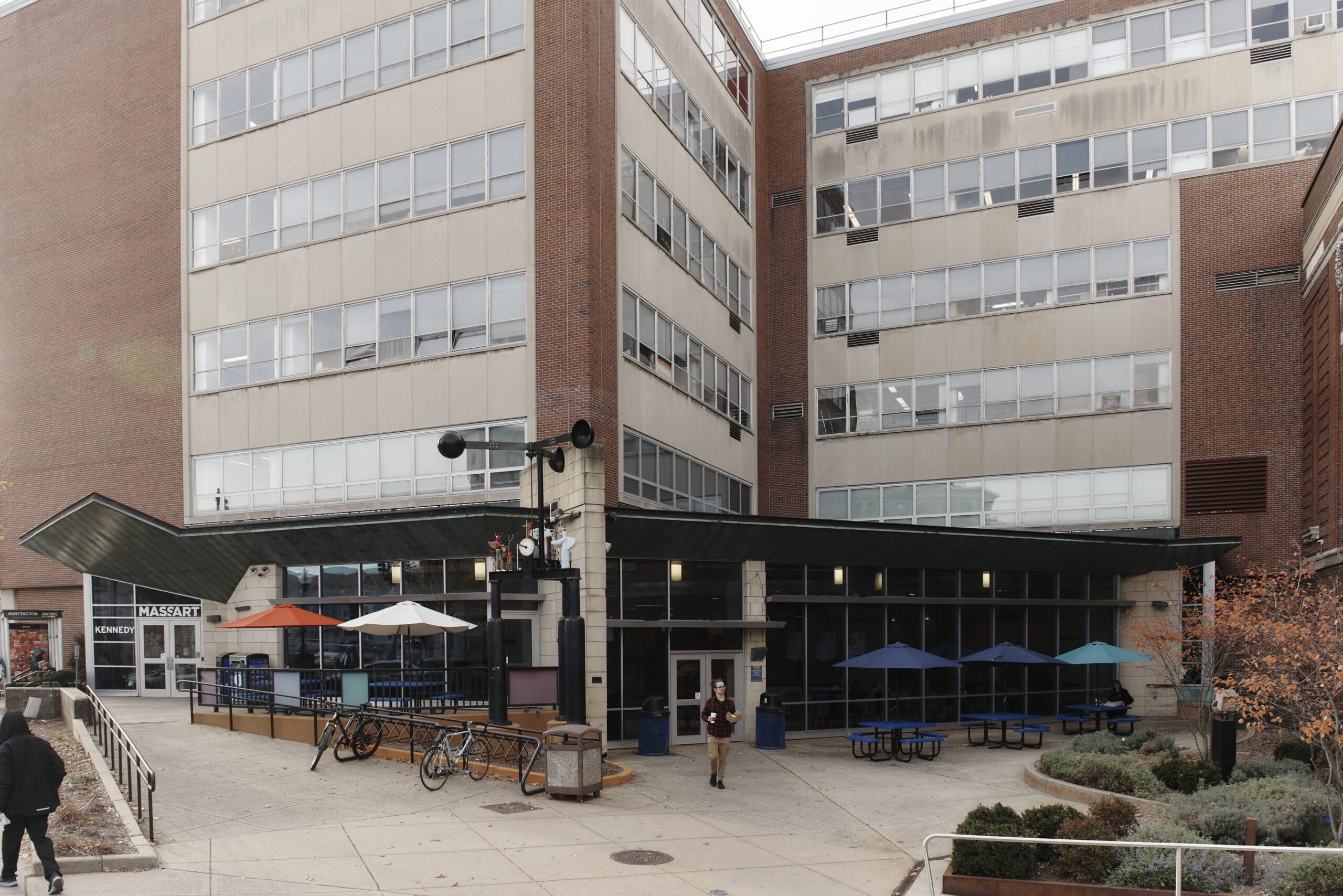 A multi-story building with a brick and glass exterior stands ready like a carefully curated portfolio. The entrance features a canopy with tables and umbrellas, perfect for preparing to enjoy the crisp air. Bicycles are parked nearby, while people walk amid the vibrant autumn foliage.