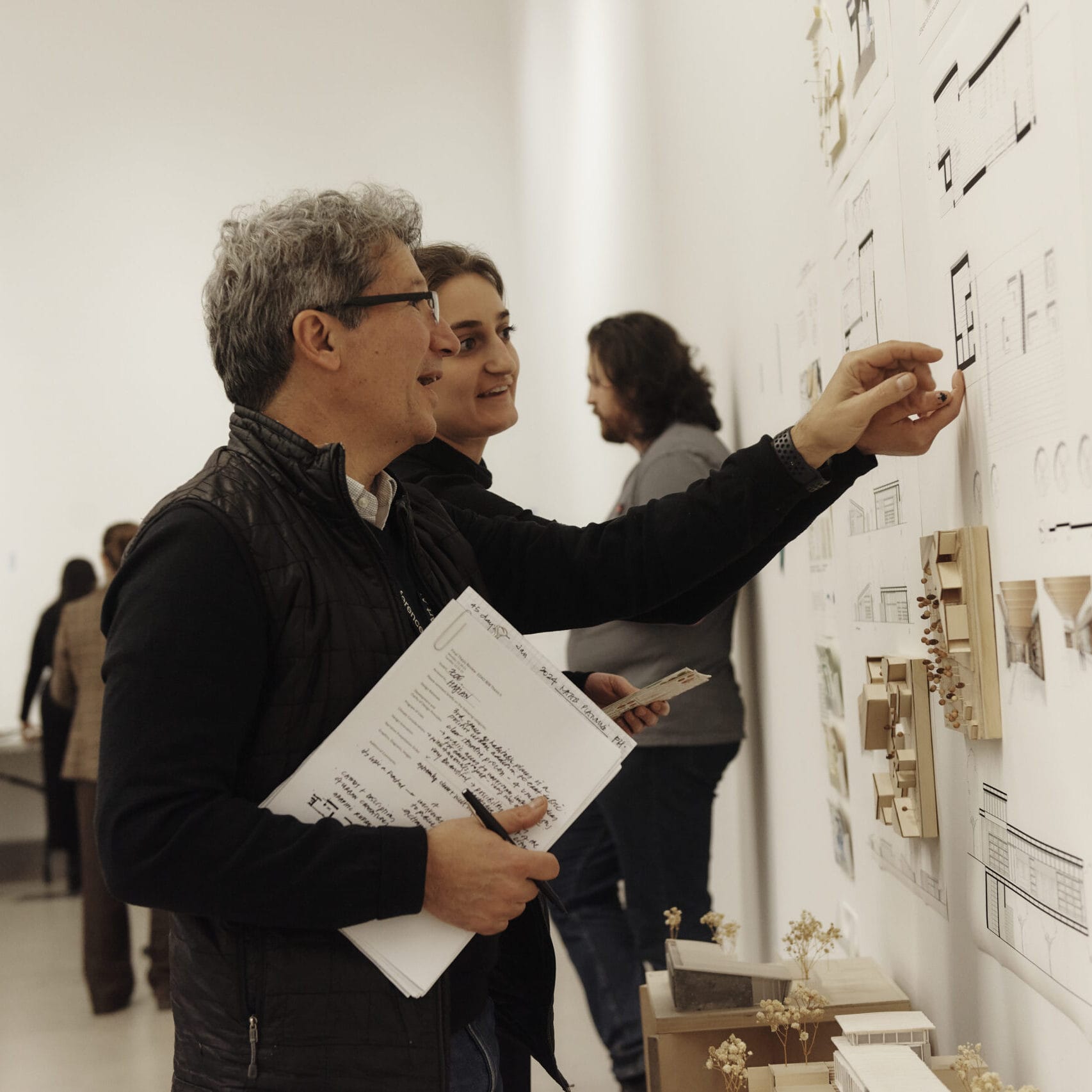 A faculty member smiles and points to a student's project on the wall, while the student looks on.