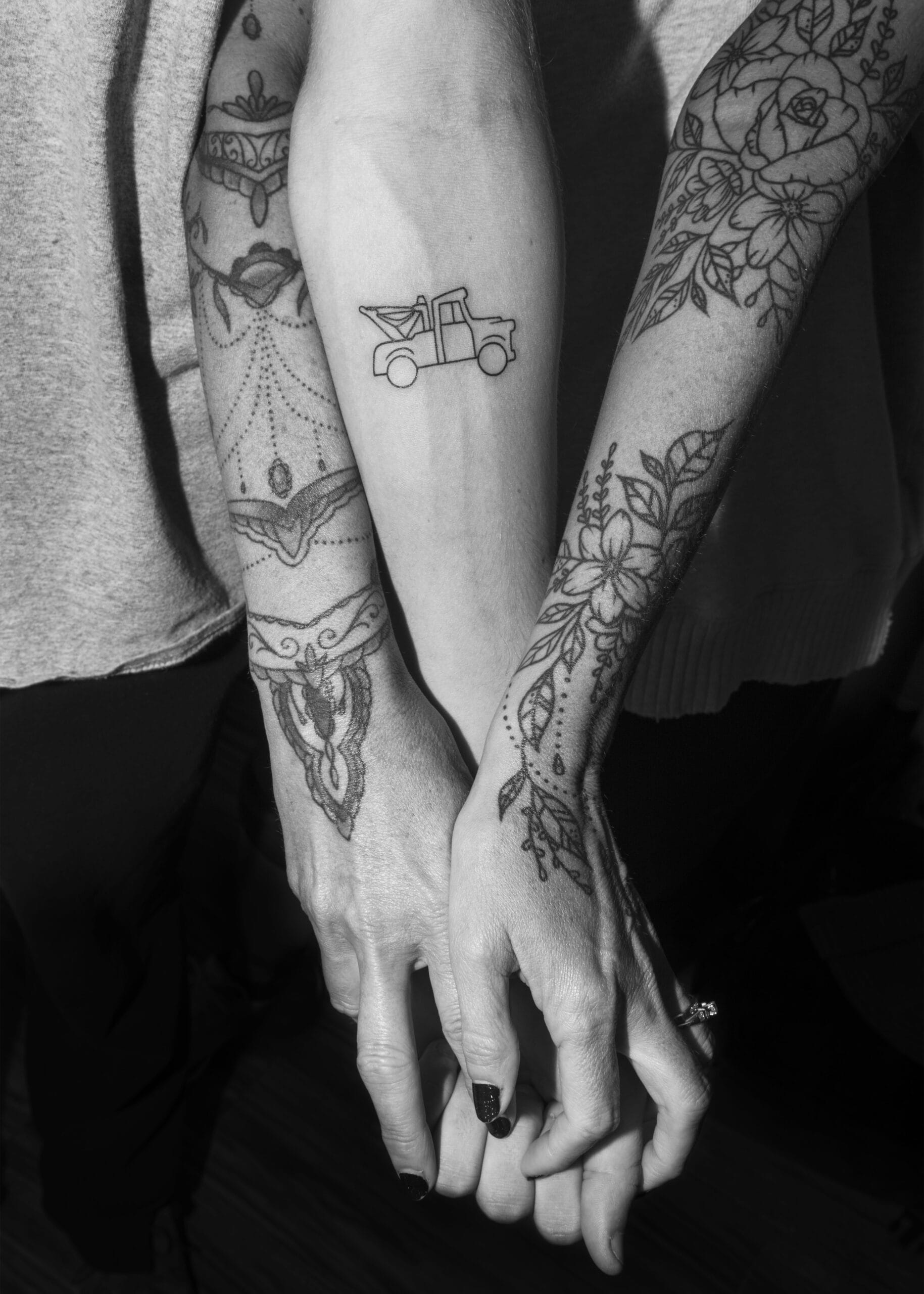 Two tattooed arms showcase a small tow truck and elegant floral patterns. Clasped hands, dressed in light shirts, suggest unity in artistry and portfolio preparation. The black and white image captures meticulous detail, perfect for any creative collection.