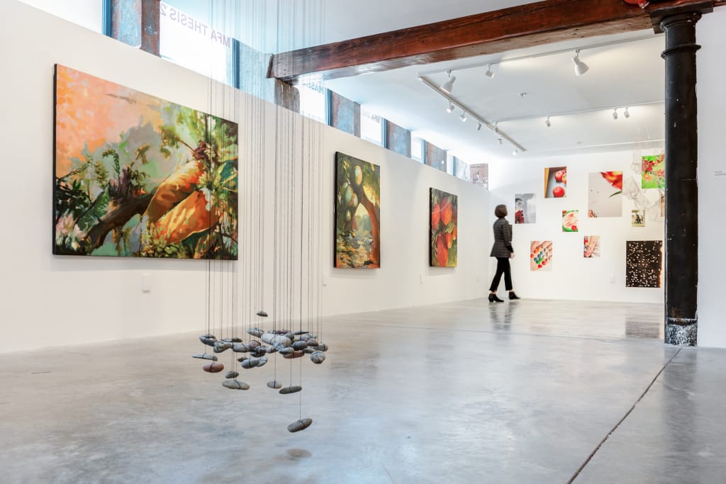 At MassArt Galleries, a person strolls through an art exhibition featuring colorful abstract paintings on white walls. In the foreground, stones hang from wires, forming a mobile-like sculpture. The space is well-lit with wood and metal beams.