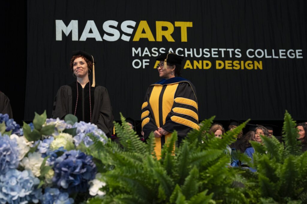 Two art leaders in academic regalia stand on stage in front of a MassArt banner. The stage is adorned with blue and white flowers and lush green ferns, embodying the spirit of creativity and innovation.