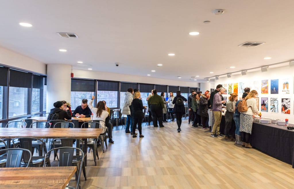 A group of people gather in a brightly lit room with large windows and wooden tables, reminiscent of MassArt galleries. Some stand in line near a table with various art items, while colorful posters adorn the walls. The atmosphere appears casual and social.
