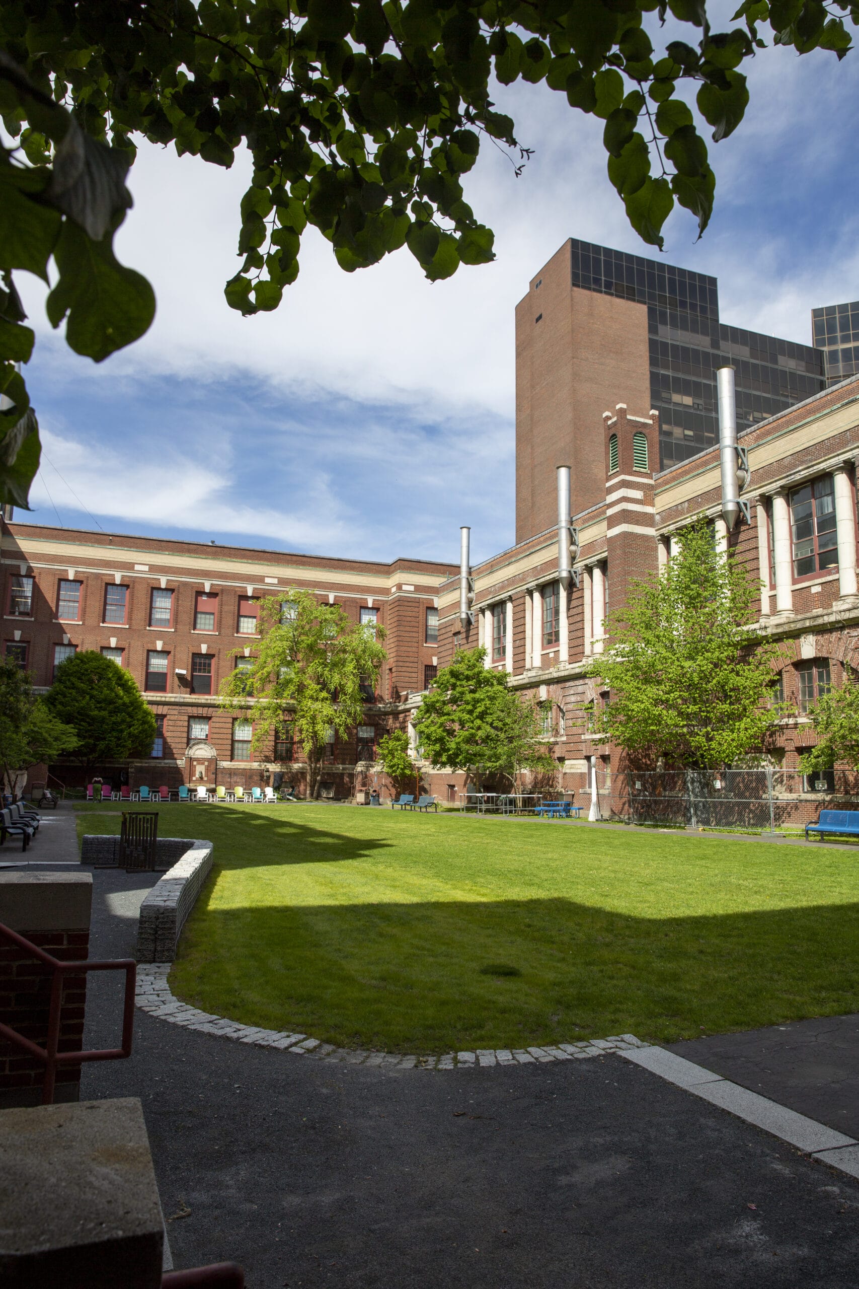 A courtyard welcoming returning students features a grassy area surrounded by red brick buildings with white-framed windows. Trees and benches line the space, while a tall building with dark windows looms in the background, set against a partly cloudy sky.