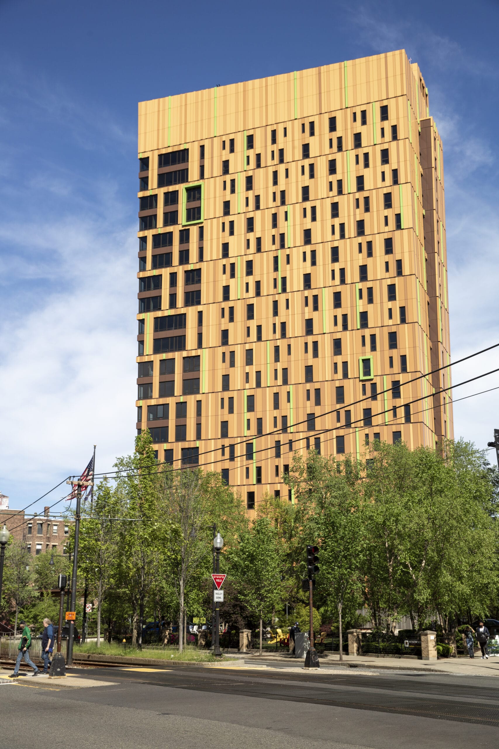 Tall, modern building with a warm, golden-brown facade and small green accents stands against a blue sky. Surrounded by lush trees, returning students walk nearby, and American flags are visible on the left. Street and traffic signs present in the foreground.