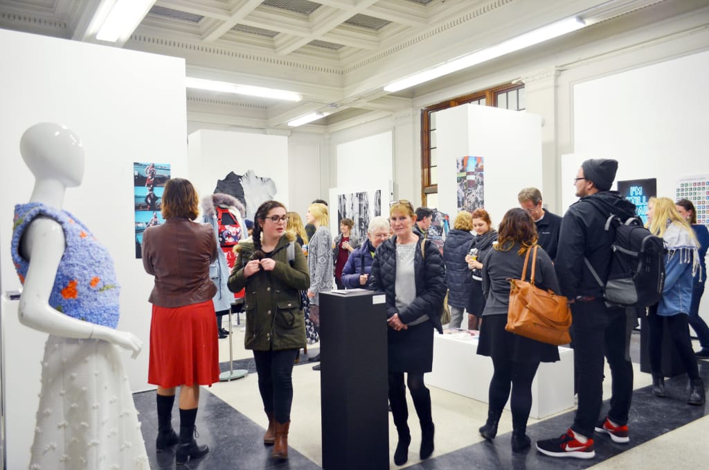 A group of people in a gallery setting from MassArt Galleries observes various artworks displayed on white walls and stands. A mannequin wearing a blue and white textured outfit stands in the foreground. The gallery, hosting one of its renowned art exhibitions, is well-lit with a modern interior.