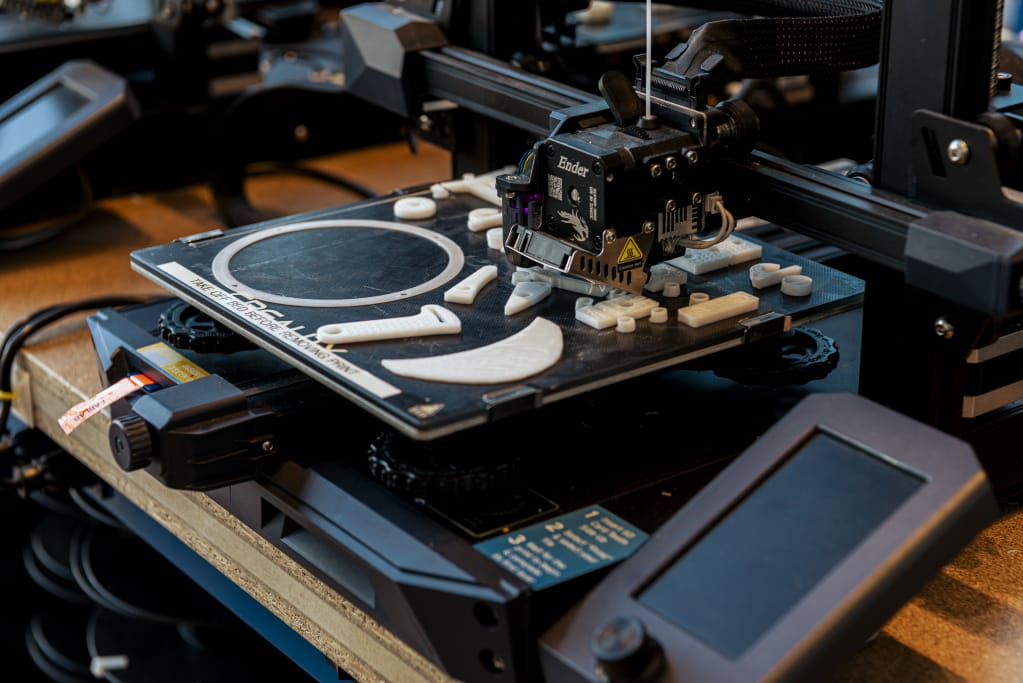In a bustling studio, a 3D printer hums in action, carefully crafting white plastic parts. Various shapes, like circular and oval pieces, take form on the print bed. The control panel and components are prominently displayed as this innovative resource brings designs to life.