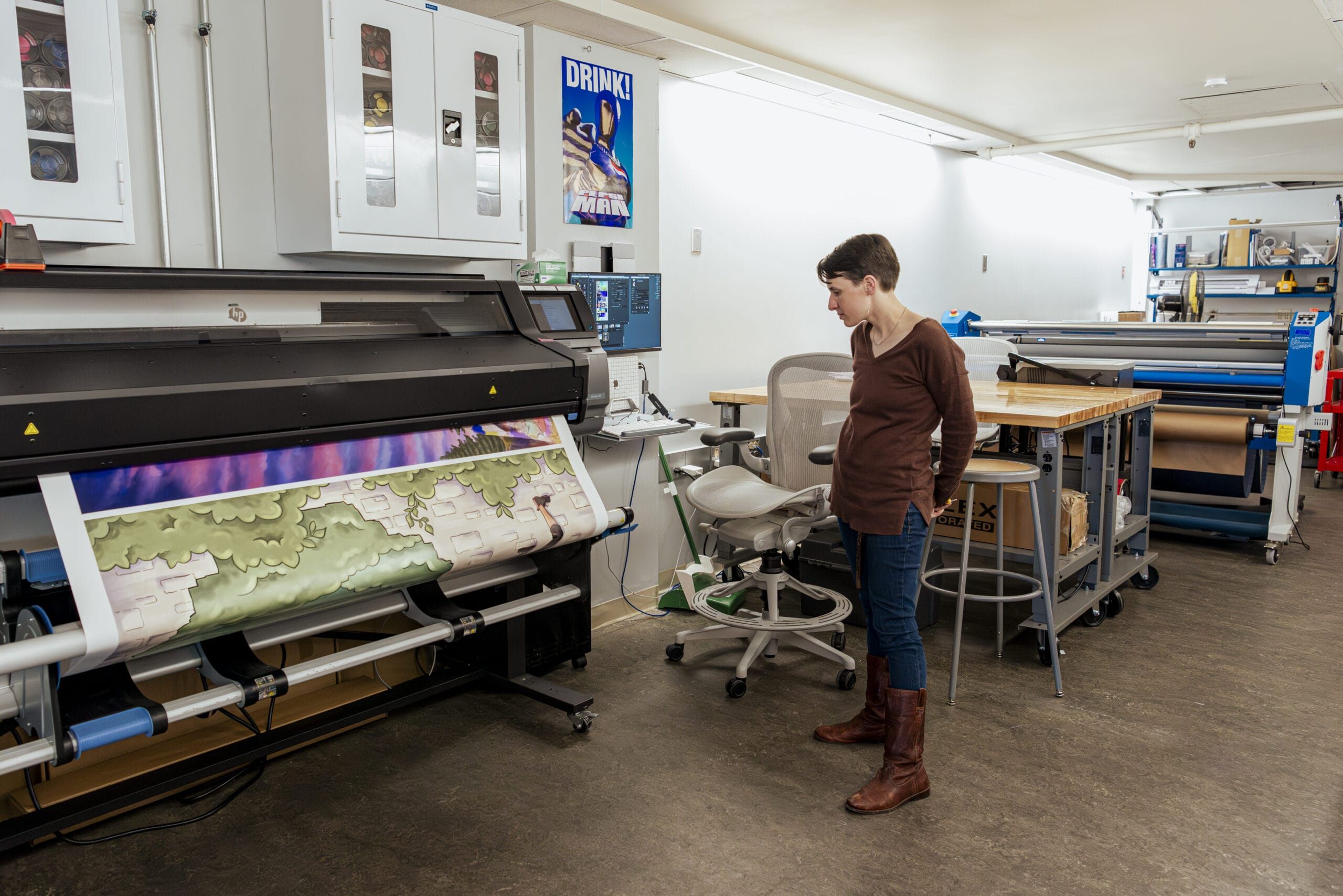 A person stands in a print shop, looking at a large printer producing a colorful landscape image. The workspace is equipped with various tools, workbenches, and storage cabinets. The setting is bright and organized.