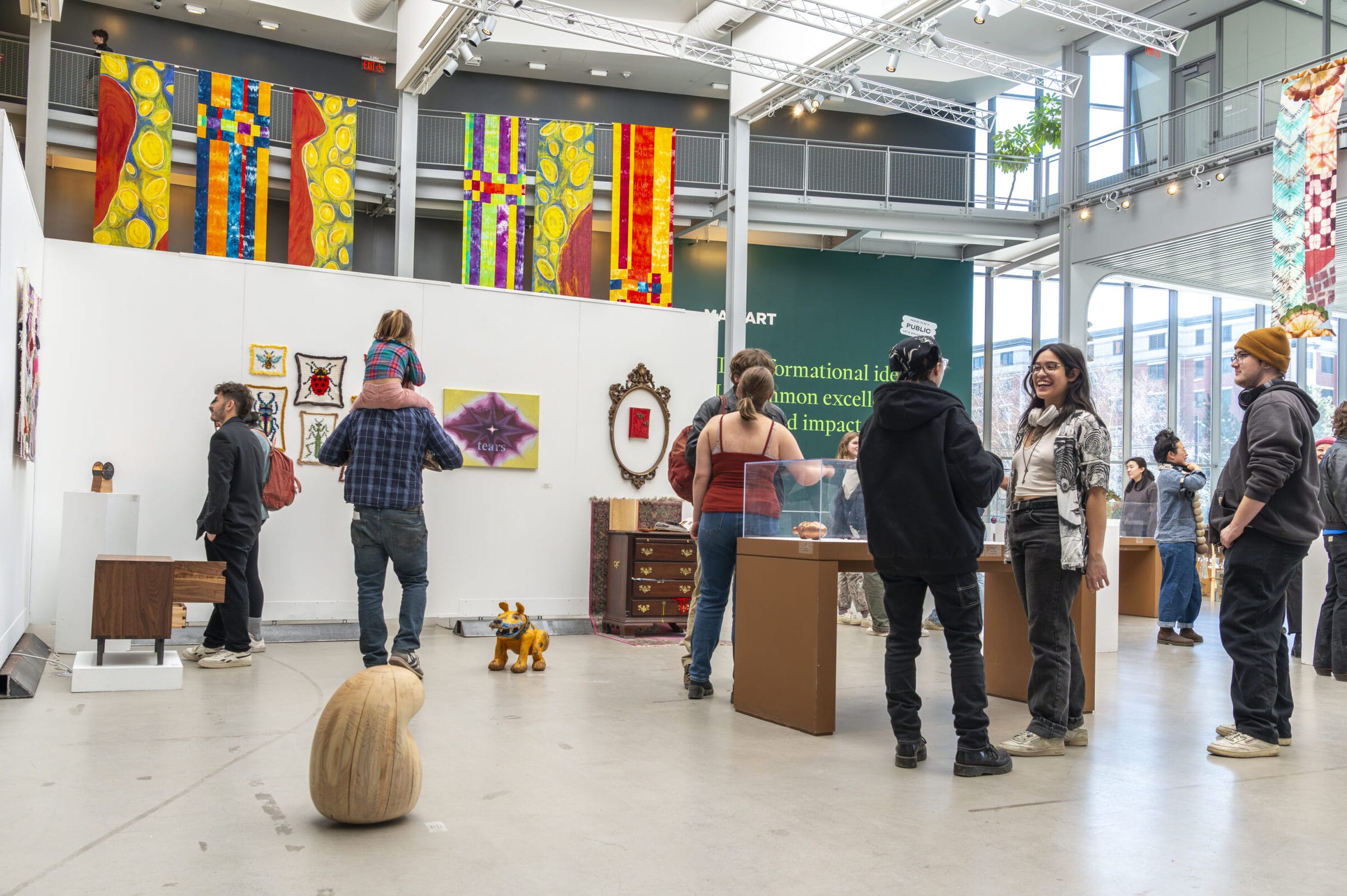 People are observing art pieces at a gallery. Colorful textile art hangs prominently above white walls. Some visitors are engaged in conversation, while others admire the diverse range of artworks on display. The setting is bright and spacious.