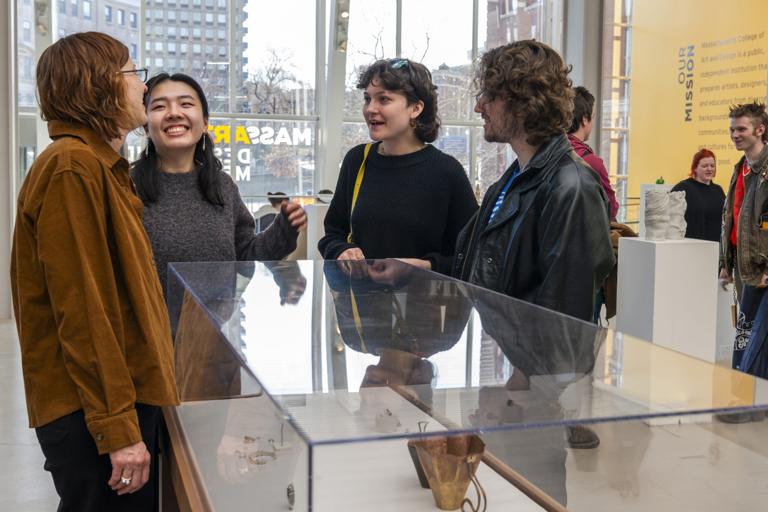 A group of graduate students gathers around a glass display case in a museum, smiling and engaging with each other. The background features large windows and a yellow wall adorned with text, creating an inviting atmosphere for their lively academic discussions.