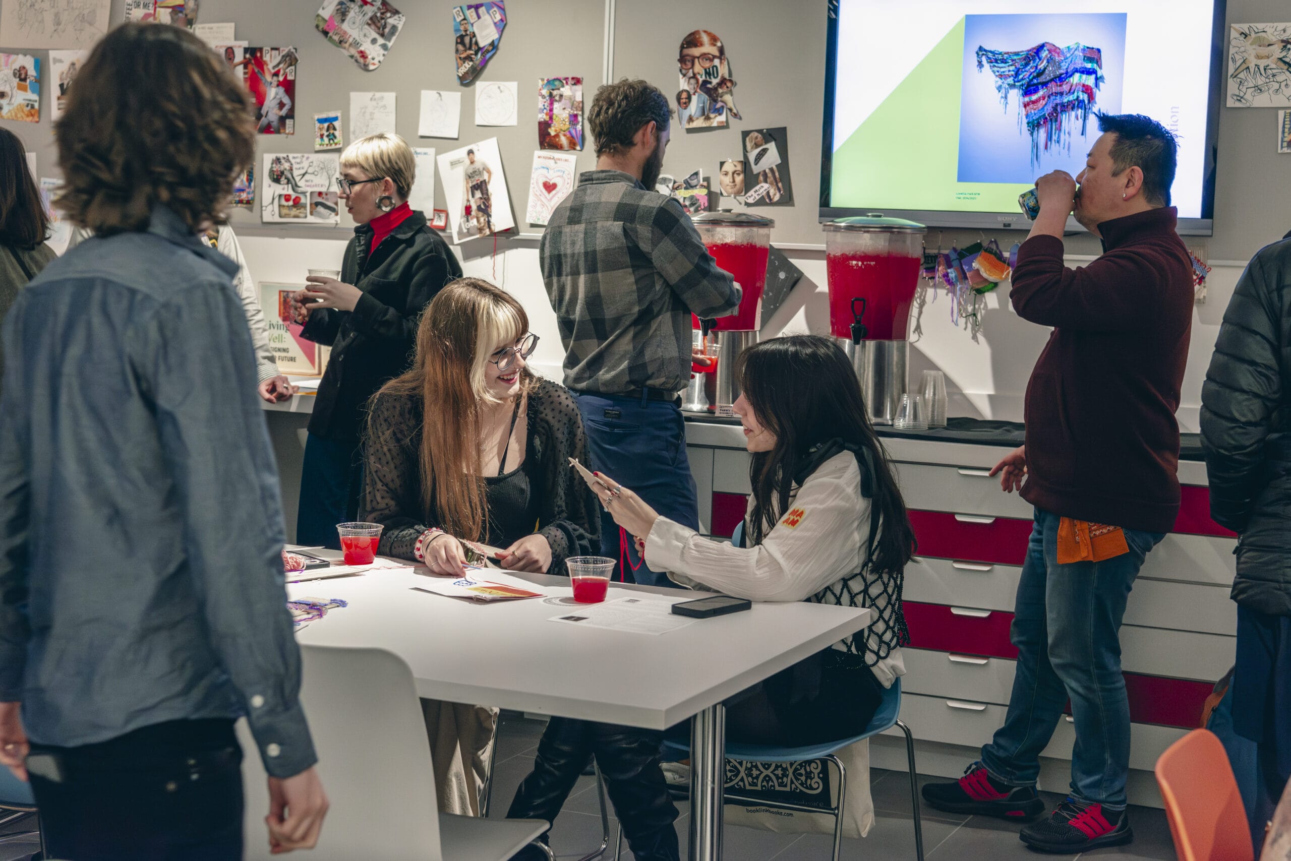 A group of people is socializing in a casual indoor setting. Some are seated at a table enjoying drinks, while others stand nearby. Art and photos adorn the walls, and a screen showcases an artwork. The atmosphere is lively and friendly, highlighting the benefits of community gatherings.