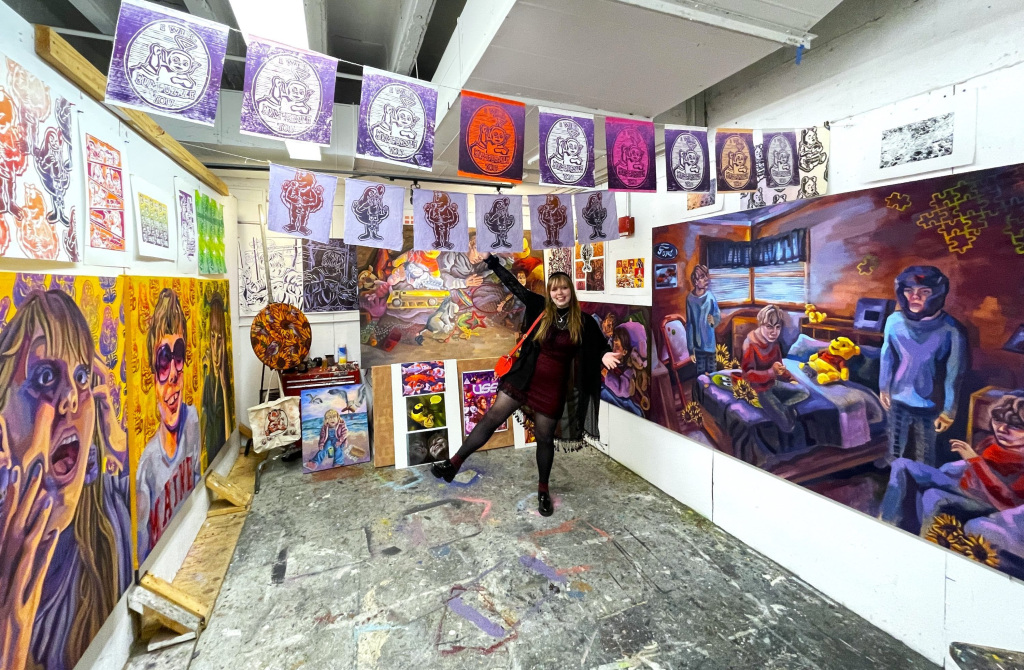 A person excitedly poses in an art studio filled with colorful, expressive paintings on the walls. Decorated banners—celebrating the 2023 Student Award winners—hang from the ceiling, enhancing the creative atmosphere, while paint-splattered floors add a vibrant touch.