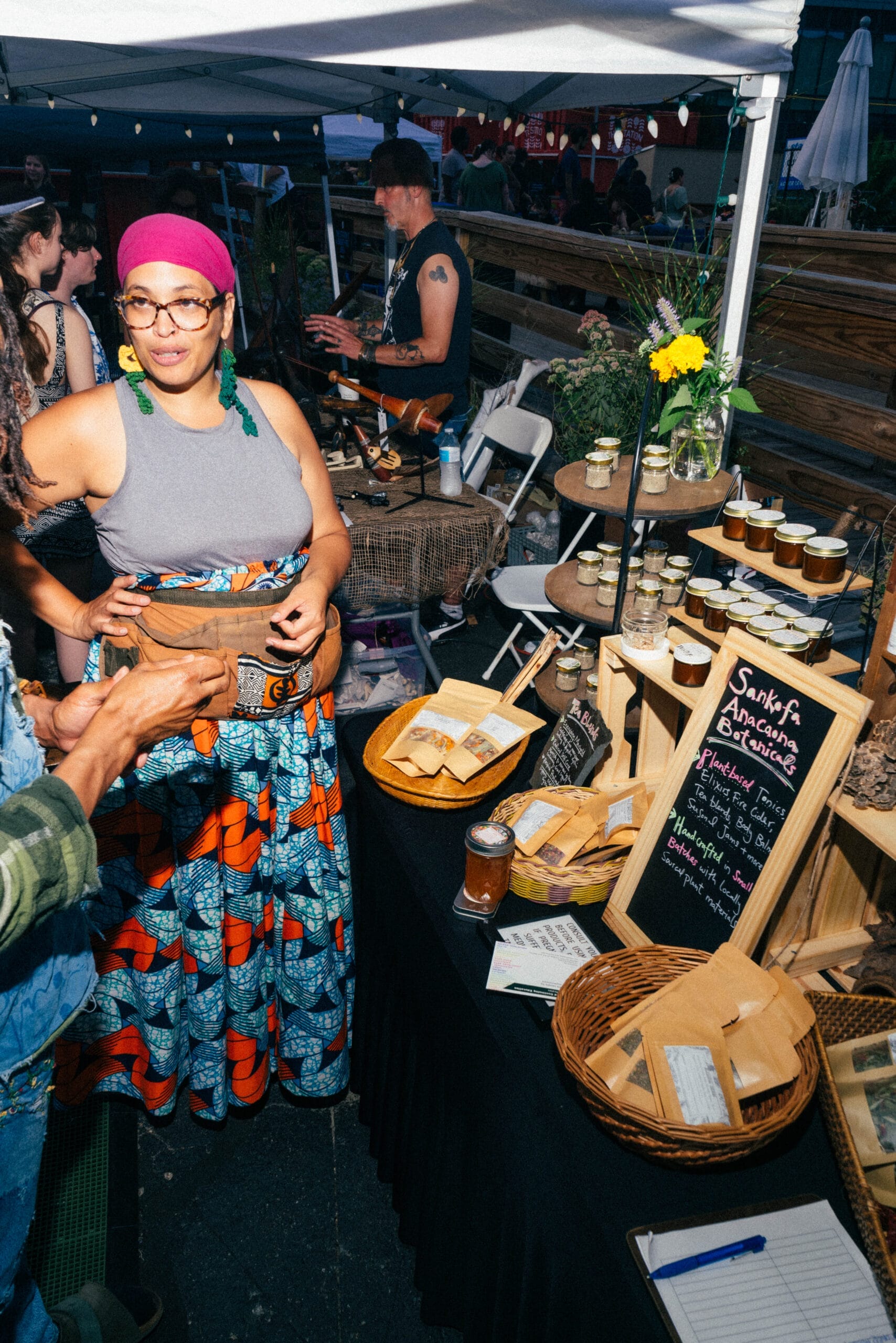 A woman in a colorful outfit stands next to a vendor table at an outdoor market, showcasing her vibrant MassArt Talent. The table brims with packaged goods and jars, accented by a chalkboard sign. People bustle in the background under a canopy, inspired to hire creatively skilled individuals.