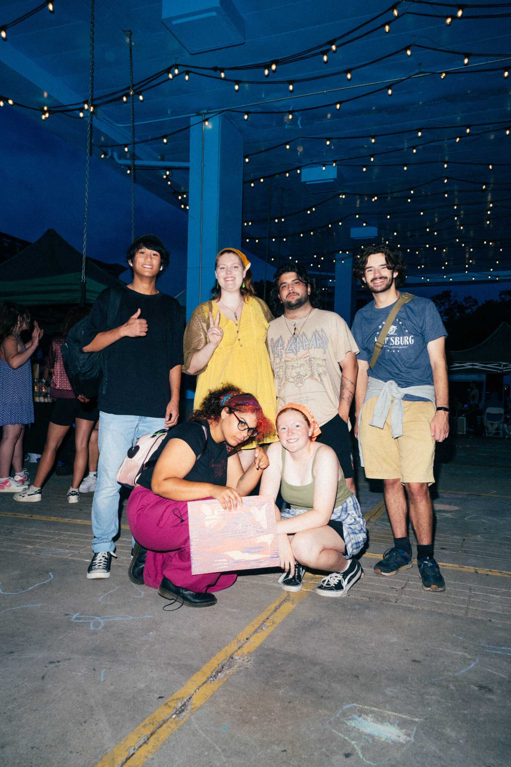 A group of six people, showcasing their MassArt Talent, poses under string lights at a lively nighttime event. One person proudly holds a large poster while others stand or crouch around them, smiling. The scene buzzes with energy as additional people mingle in the background.