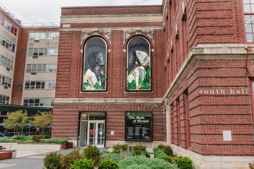 The red brick building, reminiscent of galleries, boasts large arched murals of two people gazing at each other. Below, glass doors mark the entrance, beside which a sign titled The Myth of Normal nods to MassArt’s influence. Lush greenery and a modern building complete the scene.