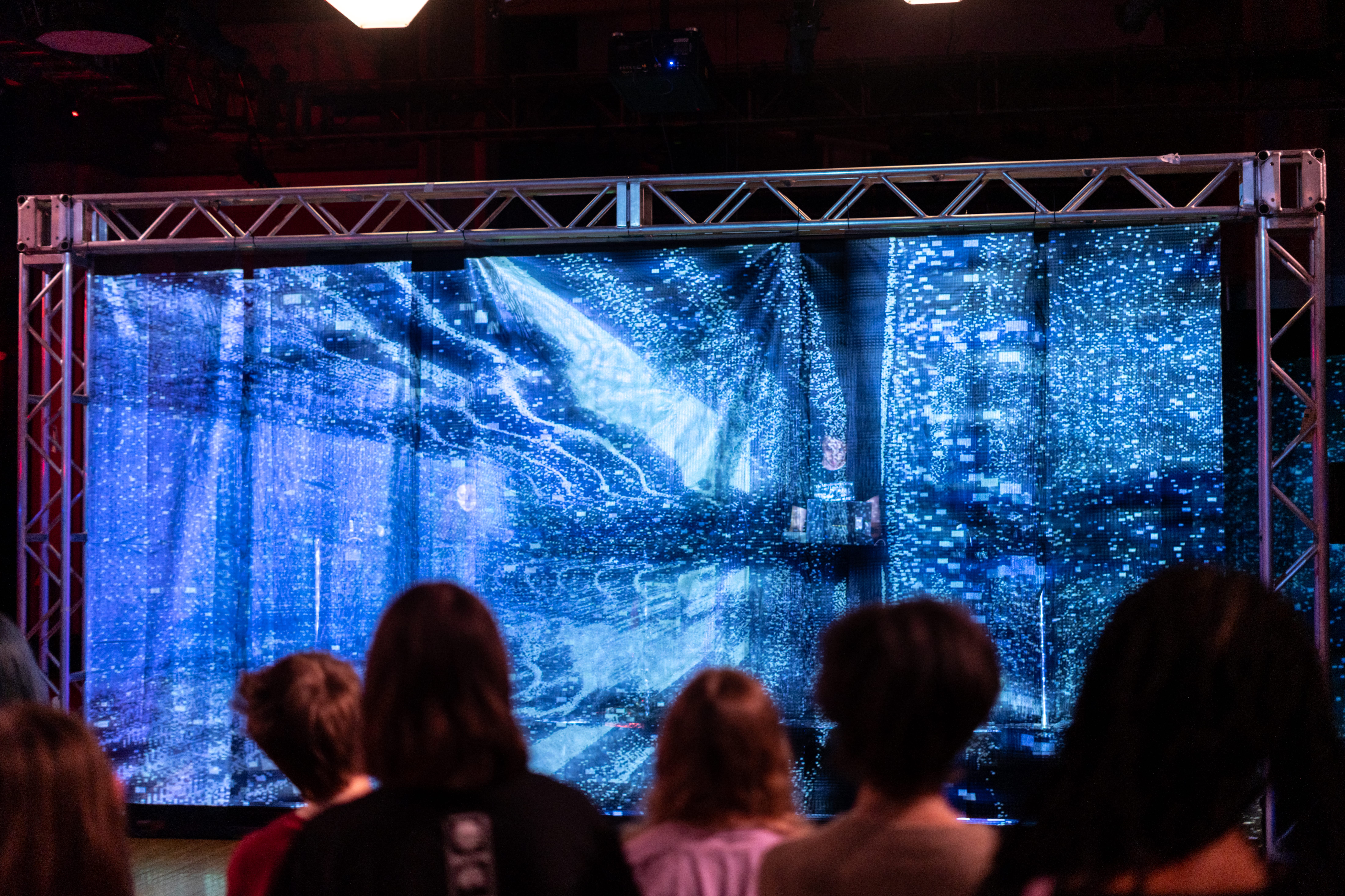 An audience watching a performance piece with blue swirly light and a figure on a stage.