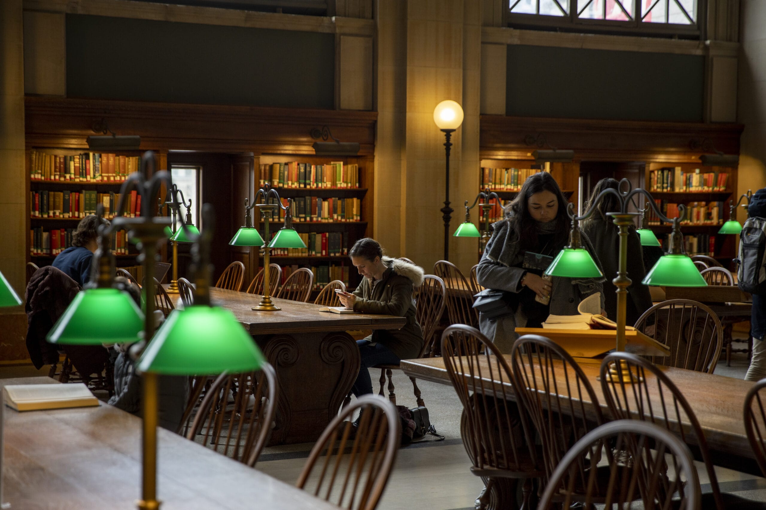 The library, ideal for cross-registration students, features wooden tables and green-shaded lamps amid towering bookshelves. A few people are reading or studying while one person searches through a bag. The room boasts a high ceiling and arched windows, offering a serene academic haven.