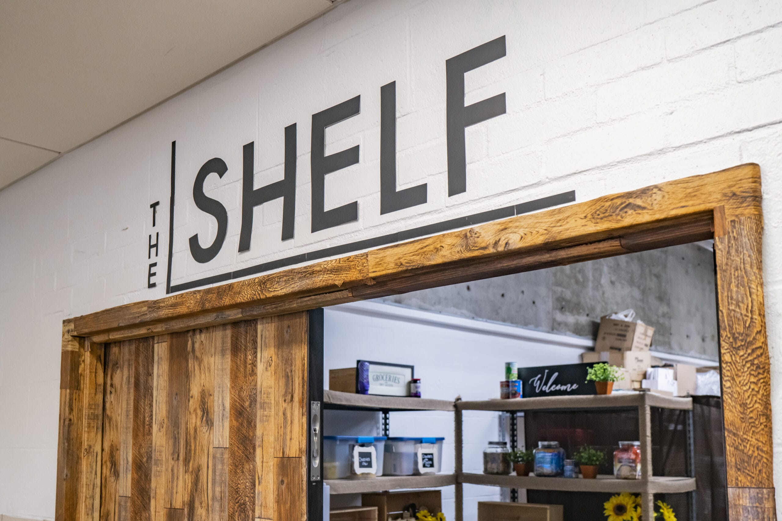 A storefront with a wooden frame features a sign that reads The Shelf. Inside, shelves display various items, including jars of food, boxes, and small decor pieces like sunflowers and a Welcome sign. The white brick wall fosters a sense of community.