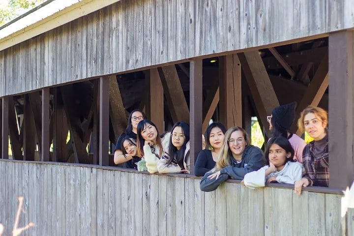 A group of people leans on the railing of a wooden covered bridge, smiling and looking towards the camera. The bridges design innovation is evident with its visible beams. Sunlight filters in, casting light across the scene, creating a perfect backdrop for memorable events.