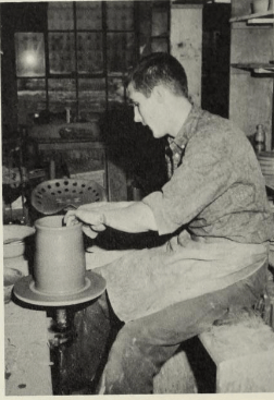 A person is shaping clay on a pottery wheel in a workshop. They are wearing a long-sleeve shirt and an apron. Shelves with pottery items and a window are visible in the background.