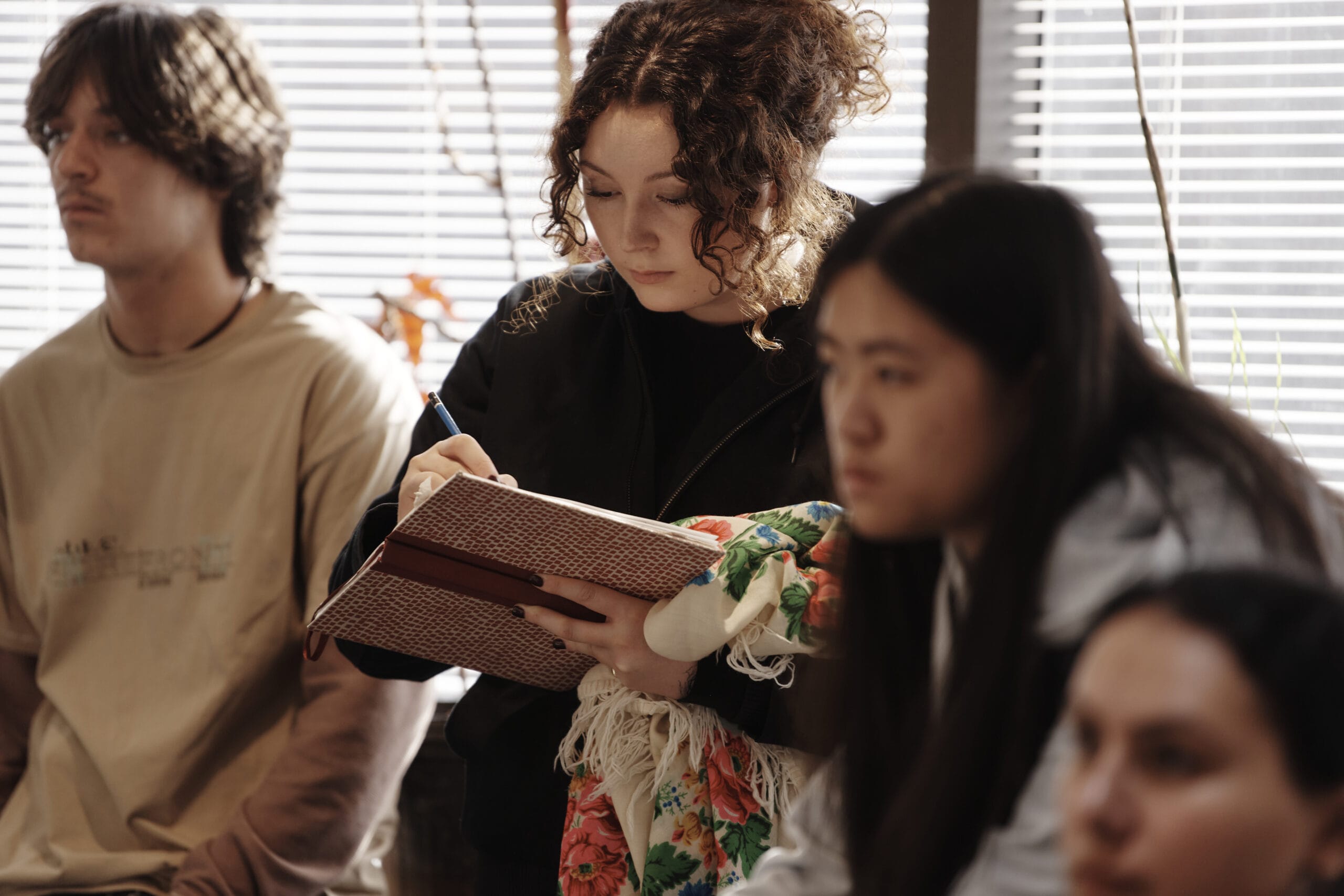 A group of students is seated in a classroom. The central focus is on a student with curly hair writing in a notebook, holding a colorful bag resting on the lap. Others appear attentive, listening, with blinds covering the windows behind them.