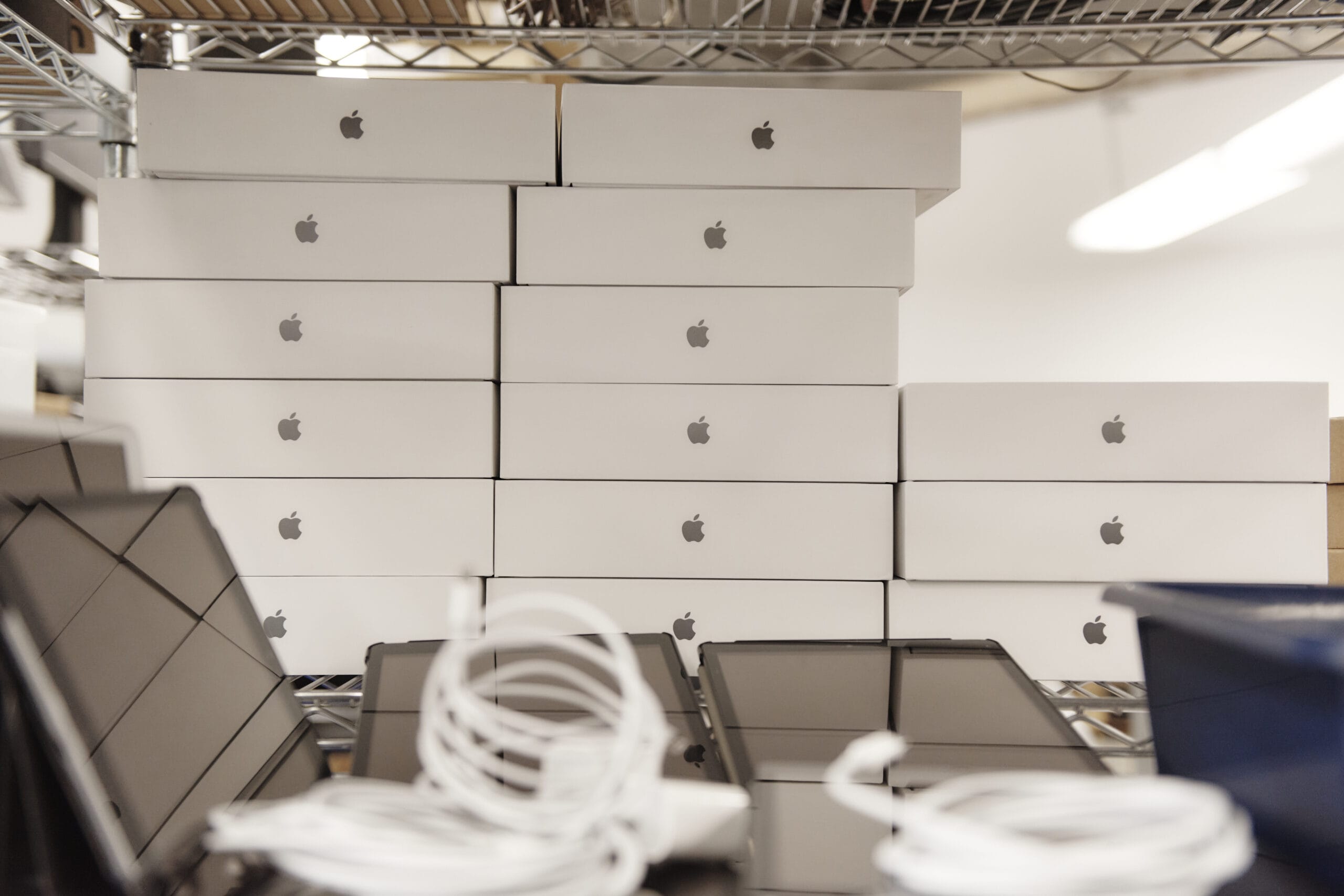 Stacked white boxes with the Apple logo are piled neatly on a metal shelf. In front, several coiled white cables and open laptops showcase cutting-edge technology, against a blurred interior backdrop.