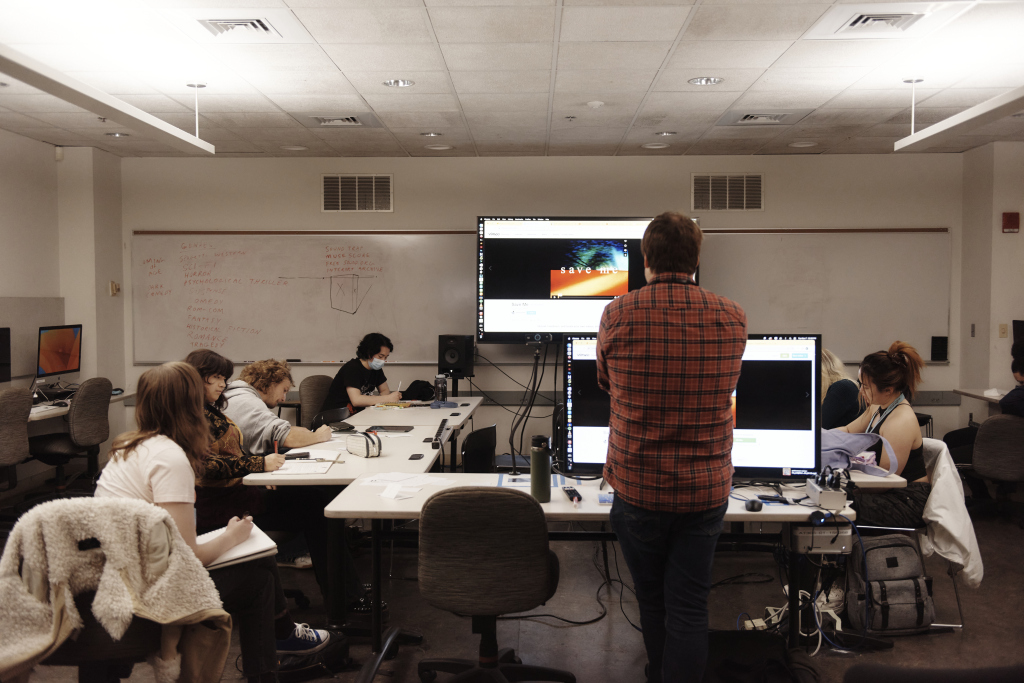 A classroom with several students working at desks features a large screen at the front displaying an image. A person stands in front of it, facing away. Whiteboards and computers are present in the room.