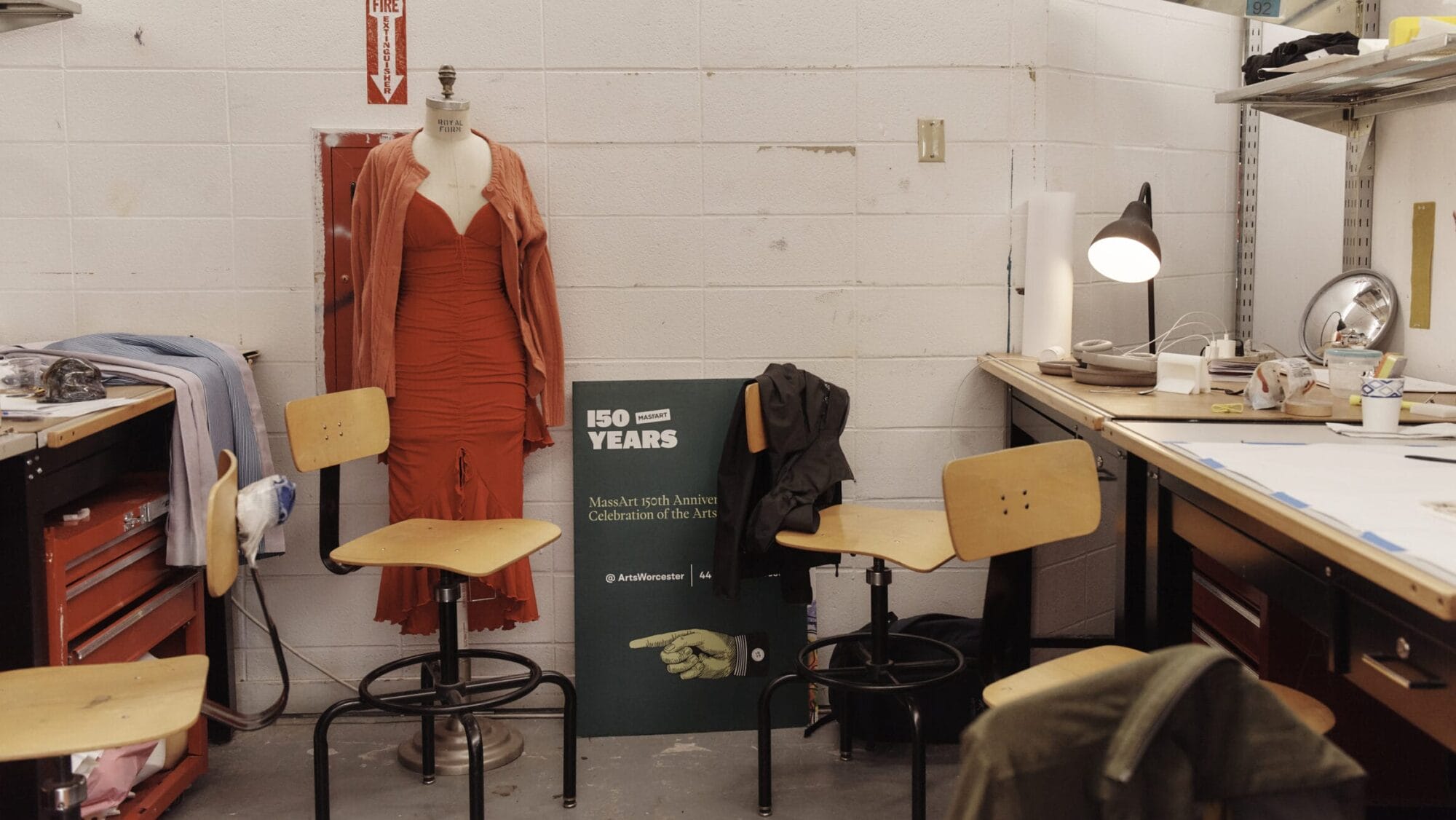 A workshop showcasing potential careers at Massachusetts College of Art and Design features a mannequin in a red dress beside a wall. Three wooden stools encircle a table cluttered with papers, topped by a lamp. A green 150 Years poster tilts against the wall, surrounded by eclectic items on tables and shelves.
